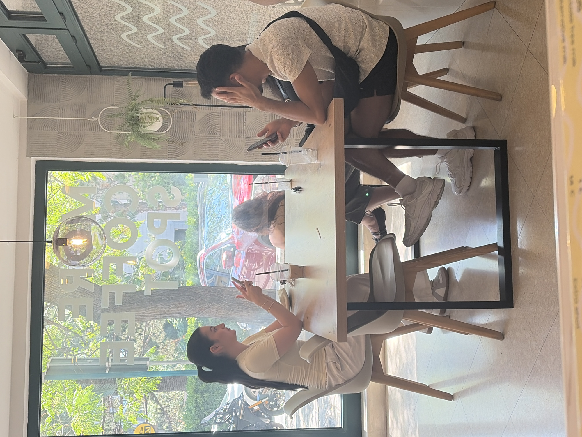 Three individuals are seated at a table in what appears to be a cafe with a large window. A woman with dark hair pulled back in a ponytail is wearing a light-colored top and pants, and is looking at her phone. A man with dark hair is wearing a white, short-sleeved shirt and dark shorts, and is holding his hand to his face while looking at his phone. A third individual, whose face is not visible, is partially visible between the two. On the table are two cups with straws, a cellphone, and a small object that could be a napkin dispenser. The cafe has textured walls and a modern light fixture hanging from the ceiling. Outside the window, trees and a red car are visible, suggesting an urban or suburban setting. The floor is tiled with a beige and white pattern. The overall impression is a casual gathering where individuals are engaged with their phones. The location is identified as Budva, Montenegro.