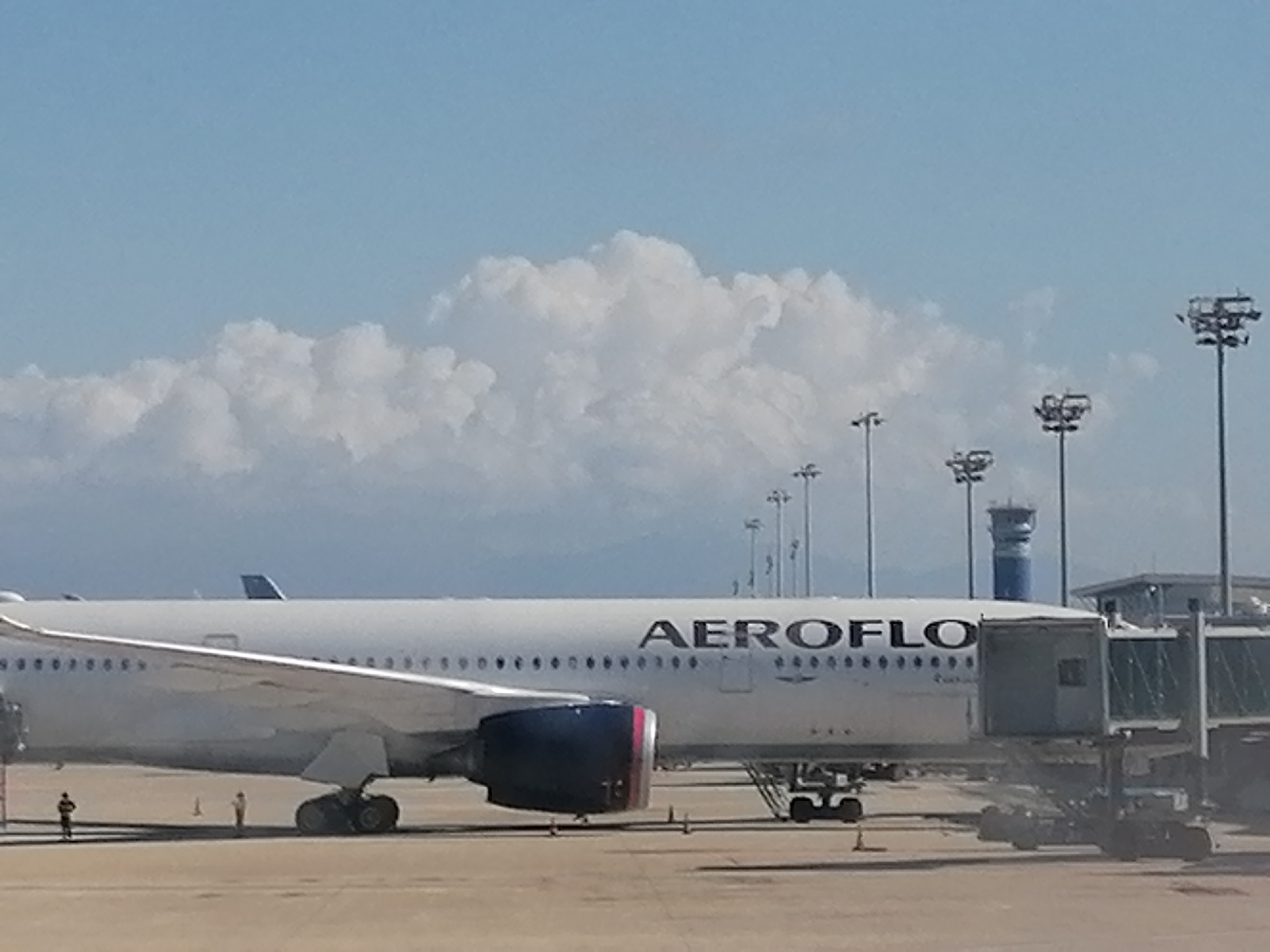 The image shows an Aeroflot airplane parked at a jet bridge at an airport. Several ground personnel stand near the plane. A control tower rises in the background, along with numerous light poles. The sky is mostly blue with cumulus clouds. The location is Unknown, Unknown.