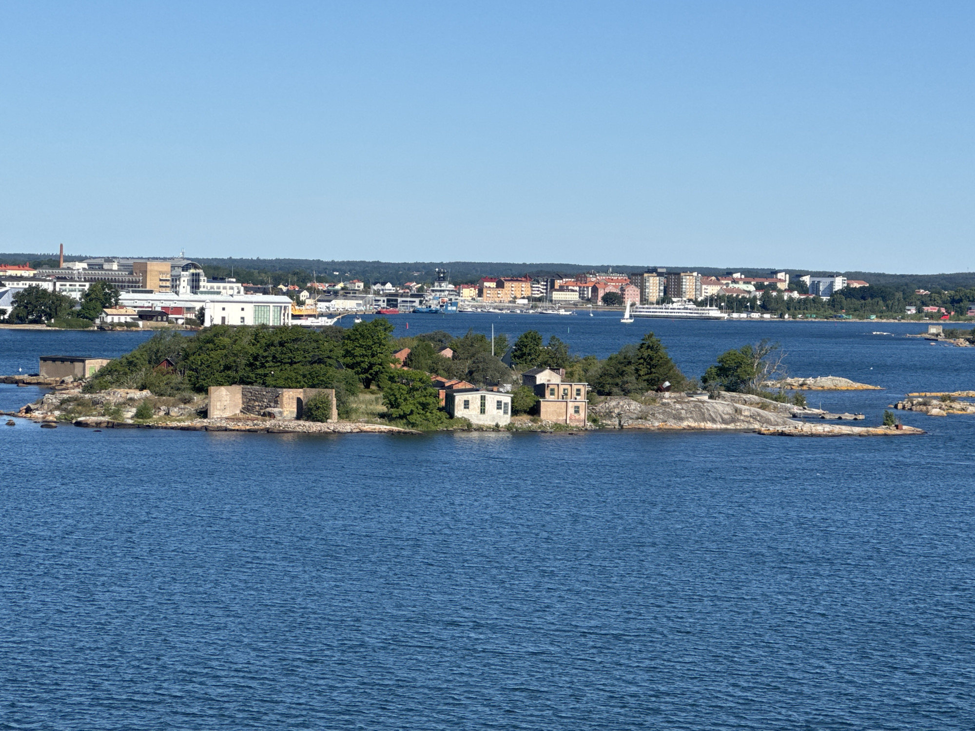 The image depicts a scenic view of the city of Karlskrona, Sweden, from the water. In the foreground, a small, rocky island is visible, dotted with a few weathered stone structures and some vegetation. Further in the background, the cityscape of Karlskrona unfolds along the coastline, characterized by a mix of modern and older buildings, including residential and commercial structures, and a harbor with various boats and ships. The water is a deep blue, reflecting the clear, bright sky. Trees and foliage are present along the shoreline and on the island. The scene appears tranquil and offers a glimpse of urban life integrated with the natural coastal environment of this historic Swedish city.