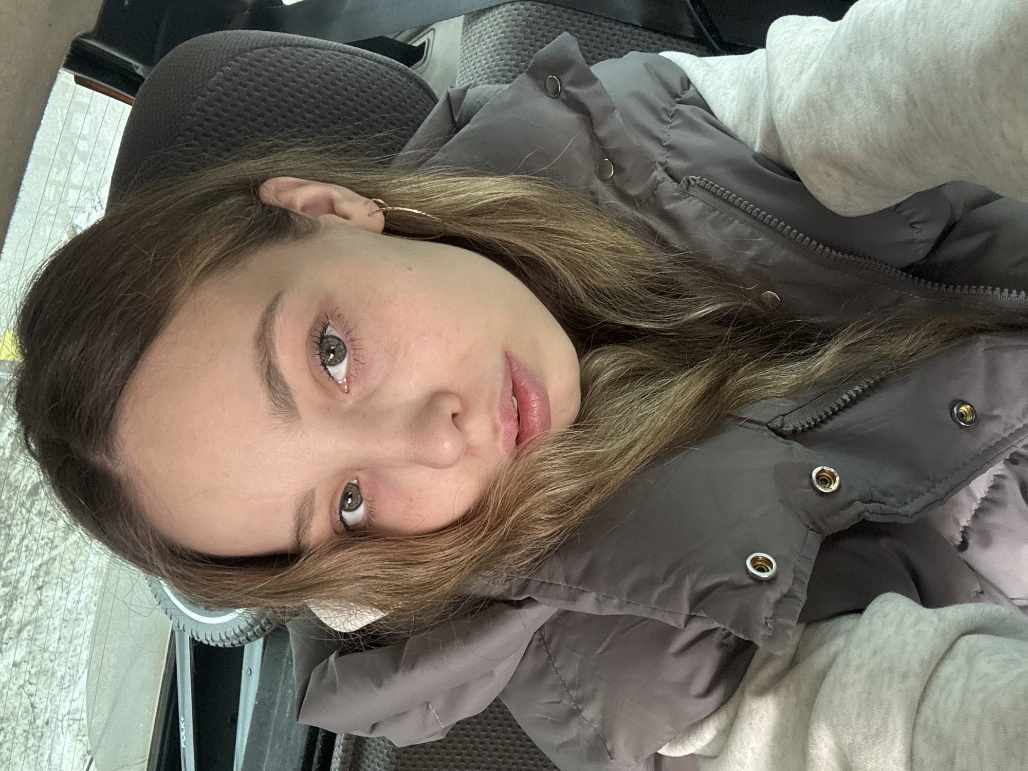 A young woman is captured in a selfie, seemingly inside a vehicle, based on the interior elements visible. She has fair skin, light brown hair with some blonde highlights, and light-colored eyes. She is wearing a grey puffer jacket over a light-colored garment, possibly a hoodie. An earring is visible in her left ear. The background includes a car seat and a glimpse of a window showing an indistinct outdoor scene. The location of Izhevsk, Russia, is not verifiable from the image.
