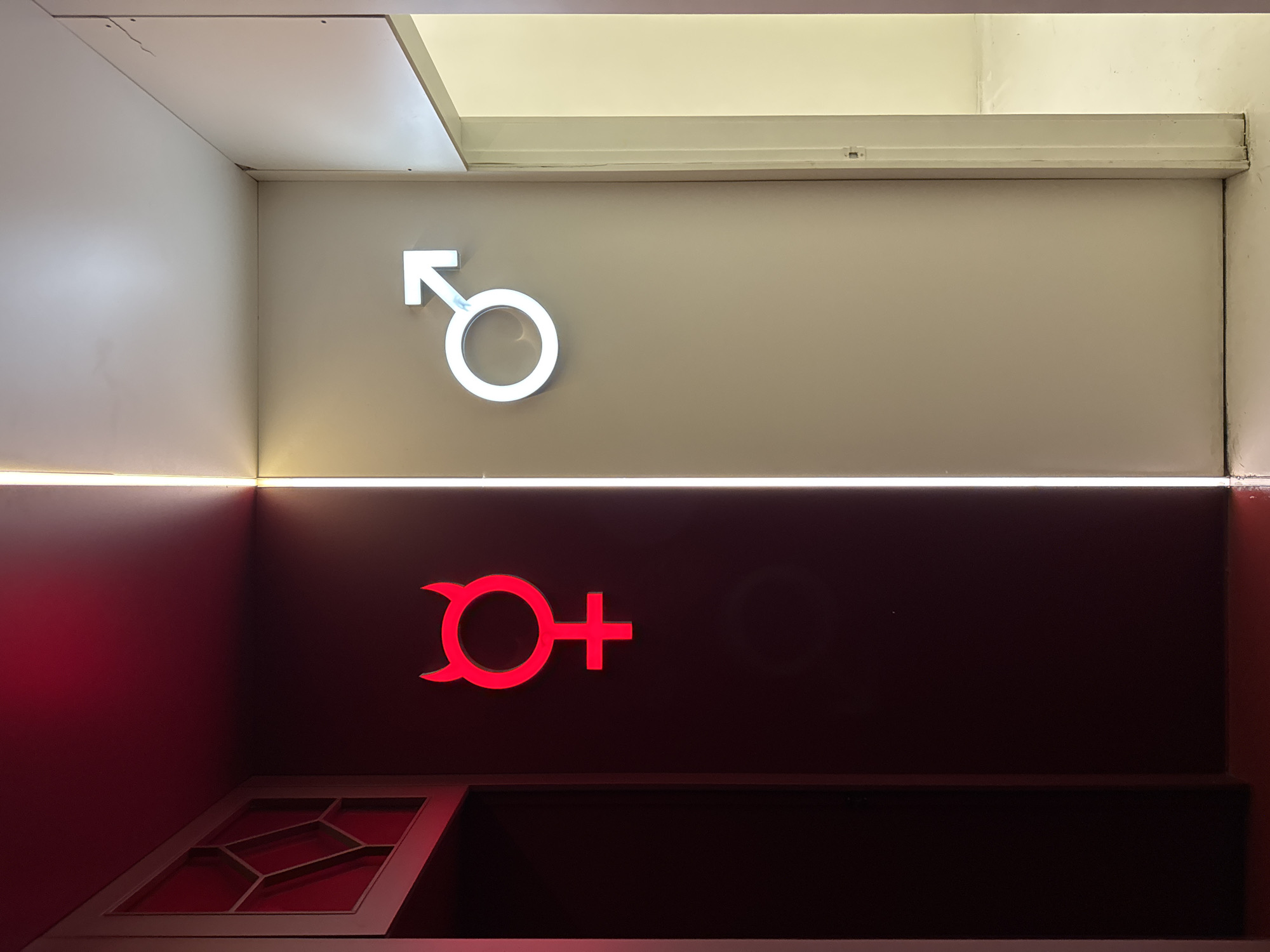 This image displays an interior wall in Sofia, Bulgaria, characterized by two distinctly illuminated symbols that suggest gender-specific facilities, possibly restrooms. The wall is divided horizontally into two contrasting sections.
The upper section features a light, neutral-toned wall, likely off-white or beige, illuminated by ambient white light from above and a horizontal strip light at its base. Centered within this section is a glowing white symbol resembling the traditional male gender sign (Mars symbol), depicted as a circle with an upward-right pointing arrow.
The lower section of the wall is a deep, dark red or maroon, also illuminated by the horizontal strip light. Here, a vibrant red symbol is prominently displayed. This symbol is a stylized rendition of the traditional female gender sign (Venus symbol), a circle with a cross below. Notably, this version is modified with two small, curved, horn-like extensions emerging from the top of the circle, adding a unique, somewhat playful or devilish character to the design.
In the bottom-left corner of the image, within the dark red area, a decorative, lighter-colored panel with a geometric, gridded pattern is visible. The strong backlighting or internal illumination of the symbols, combined with the contrasting wall colors and strip lighting, creates a striking and modern visual effect. The scene suggests a contemporary indoor setting, possibly in a commercial establishment or entertainment venue. There is no visible text, only the two distinct symbols.