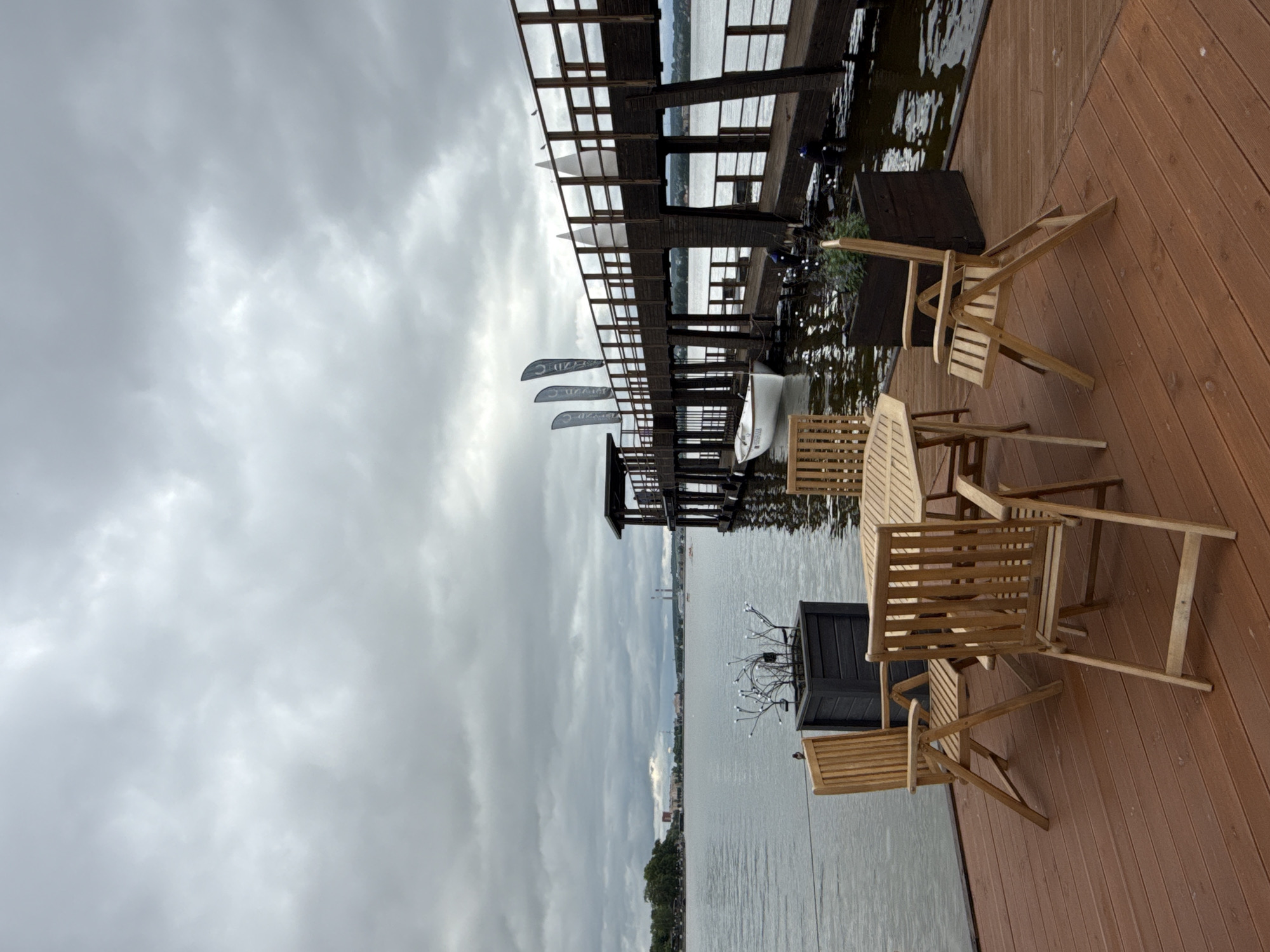 The image captures a tranquil outdoor scene on an overcast day in the city of Votkinsk, Russia, overlooking a large body of water, likely the Votkinsk Reservoir. In the foreground, a brown wooden deck features an octagonal wooden table surrounded by four foldable wooden chairs, three upright and one folded and leaning. Two dark grey rectangular planters are present: the left one contains decorative elements resembling white illuminated branches, and a small brown bird is perched on a wire extending from it towards the water; the right planter holds green foliage.
Extending from the deck into the water is a substantial dark brown wooden pier or walkway, complete with railings. A small, white open boat is moored alongside the pier near the deck. Further along the pier, three dark vertical banners with white markings are affixed to the railing, along with several white conical or sail-shaped decorative structures. Two small birds are perched on the far railing of the pier. The sky is completely covered by thick, grey clouds, indicating an overcast condition. The water surface is calm with slight ripples, reflecting the dull sky. In the distant background across the water, a shoreline with trees and indistinct city buildings is visible, including two prominent industrial chimneys or towers on the horizon. No people are discernible in the scene.