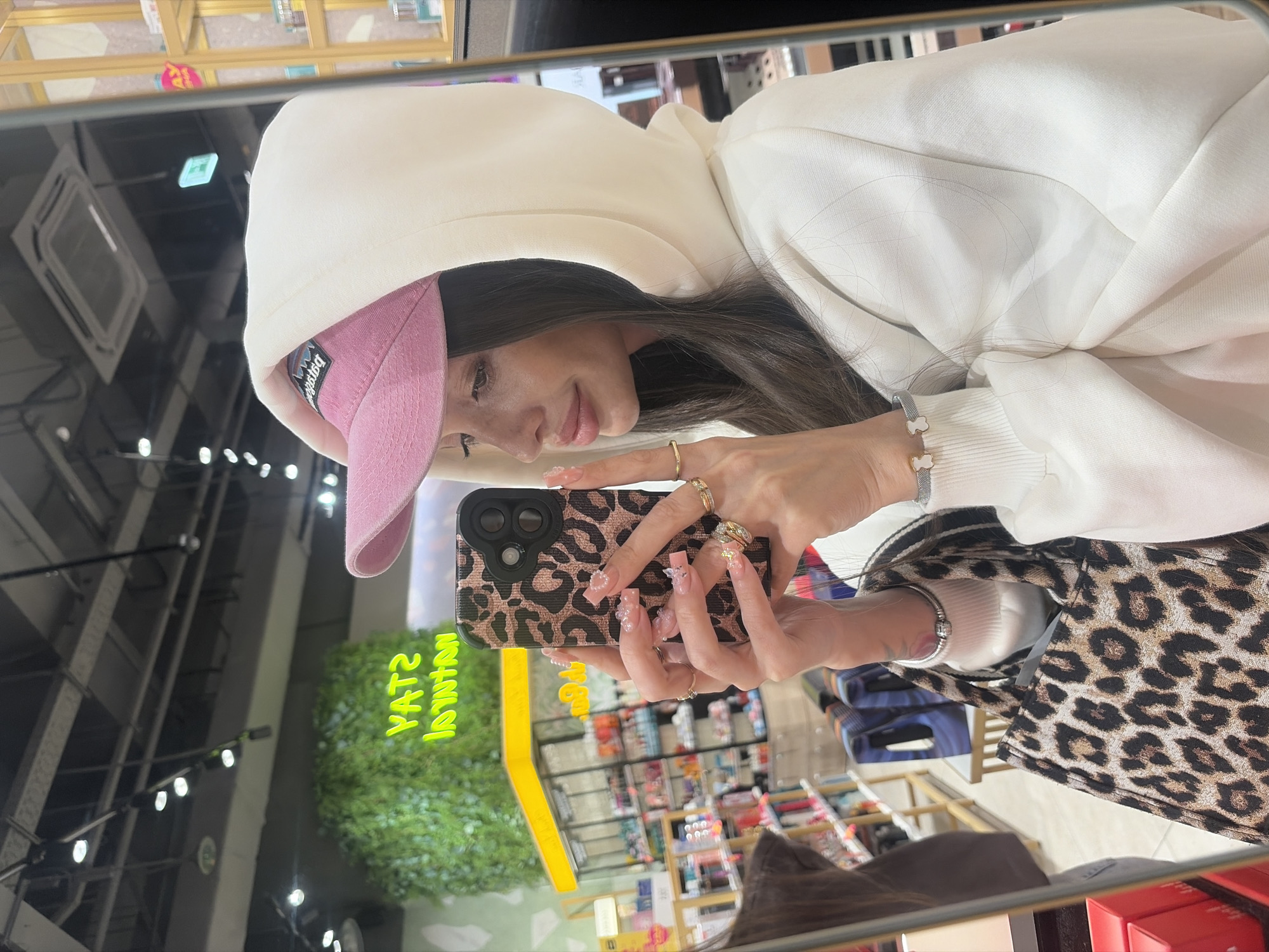 A young woman is captured in a mirror selfie, reflecting an indoor commercial setting, likely in a store or shopping mall in Izhevsk, Russia. She is wearing a white hooded sweatshirt and a pink baseball cap, which has a reversed "Patagonia" logo visible. Her long, dark hair cascades over her shoulders. She holds a smartphone with a distinctive leopard print case and a visible triple camera setup, indicating she is actively taking the photo.
Her hands are adorned with multiple rings on several fingers, and she sports intricate, long manicured nails with pink, clear, and pearlescent designs, including small bows and pearls. On her left wrist, she wears a bracelet featuring two white bear or flower-shaped charms, while her right wrist has a simpler silver bangle. A glimpse of a leopard print bag or jacket strap is visible on her right shoulder.
The background reflected in the mirror reveals store shelves filled with various products, suggesting a retail environment. Overhead lighting fixtures and ceiling vents are also visible. A large, vibrant green artificial plant wall with a neon-like yellow sign reflecting green text—partially readable as "YAT2 YAL" (likely "STAY REAL" reversed)—adds to the modern store ambiance. The woman has a calm and slightly smiling expression.
