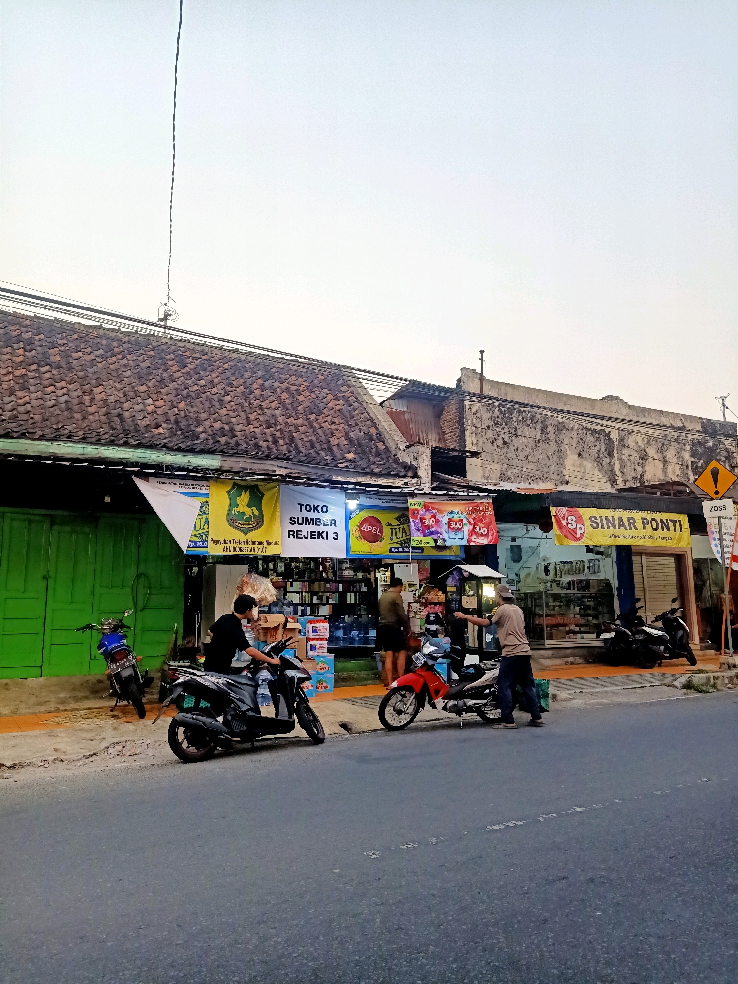 The image shows a street scene in Klaten, Indonesia. In the foreground is a grey asphalt road. On the right side of the road, there is a sidewalk. Behind the sidewalk, there are two shops with signs. The shop on the left has a green door and is displaying banners and products. The shop on the right has a yellow banner with "SINAR PONTI" written on it. There are several motorcycles parked on the sidewalk and in front of the shops. Two men are standing near a red motorcycle in front of the shop on the right, and one man is on a black motorcycle in front of the shop on the left. The buildings have tiled roofs and weathered walls. The sky is overcast.