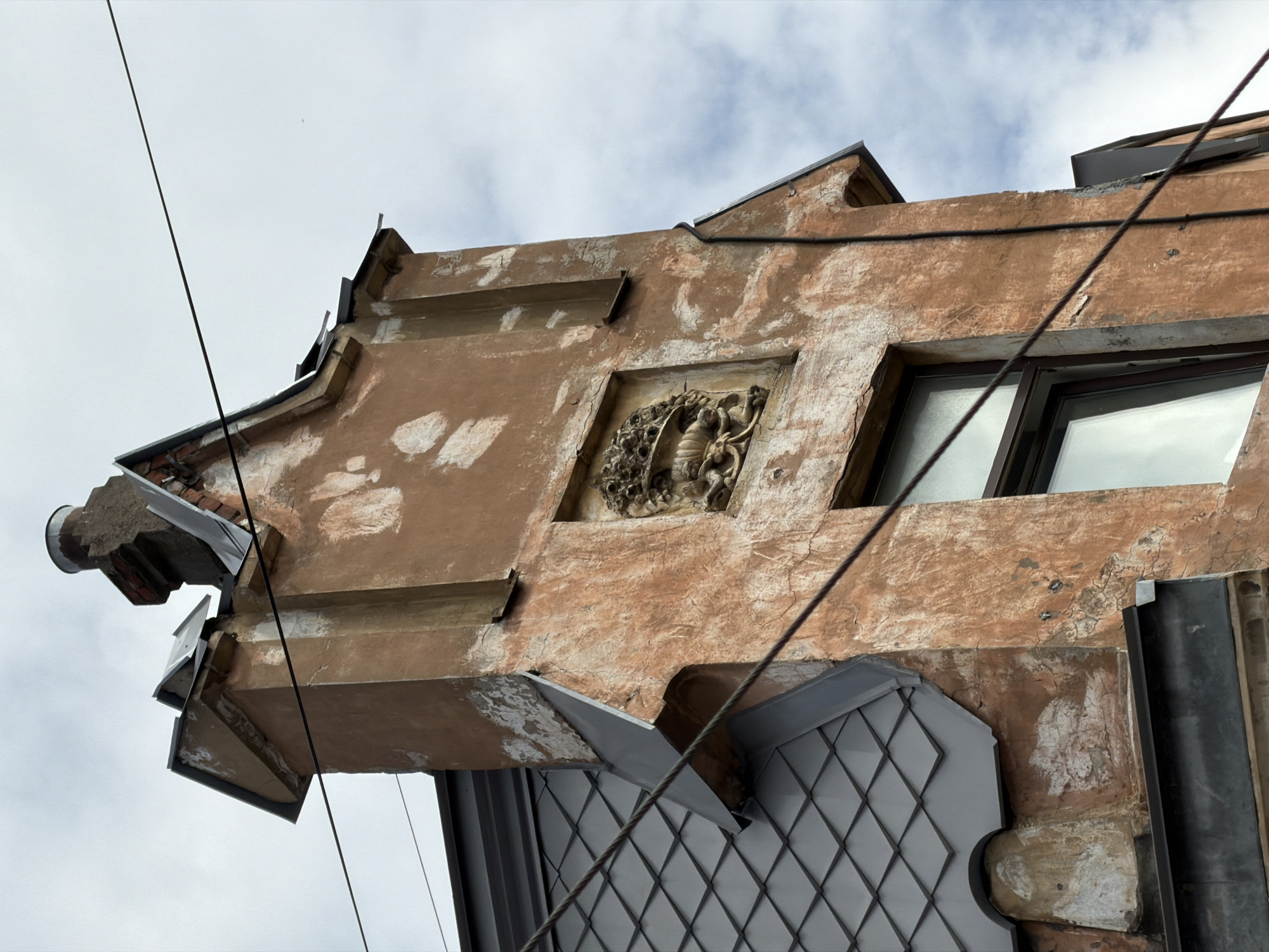 The media presents an upward-angled view of an ornate, aged building facade in Saint Petersburg, Russia, set against an overcast, cloudy sky. The building's exterior is a weathered reddish-brown or terracotta, with numerous patches of lighter, possibly original, plaster showing through, indicating significant wear and historical character.
A prominent feature is a central, square bas-relief carving depicting an intricate, grotesque, or mythical figure, possibly a dragon or serpent, intertwined with decorative scrollwork. To the right of this carving, two windows with dark frames are visible, their panes reflecting the cloudy sky. The upper section of the facade suggests a gabled roofline or an elaborate cornice, accented with dark grey or black metal flashing.
On the far left, a functional, stone-like water spout or drainage element protrudes from the wall. Below the main facade, a lower section, possibly a bay window or a detailed part of the roof, is clad in dark grey metal sheets arranged in a diamond or grid pattern. A thick, dark cable runs diagonally across the image, partially obscuring the architecture, and a thinner cable runs horizontally along the upper wall. The scene suggests a daytime setting with diffused light. No visible text is present in the image.