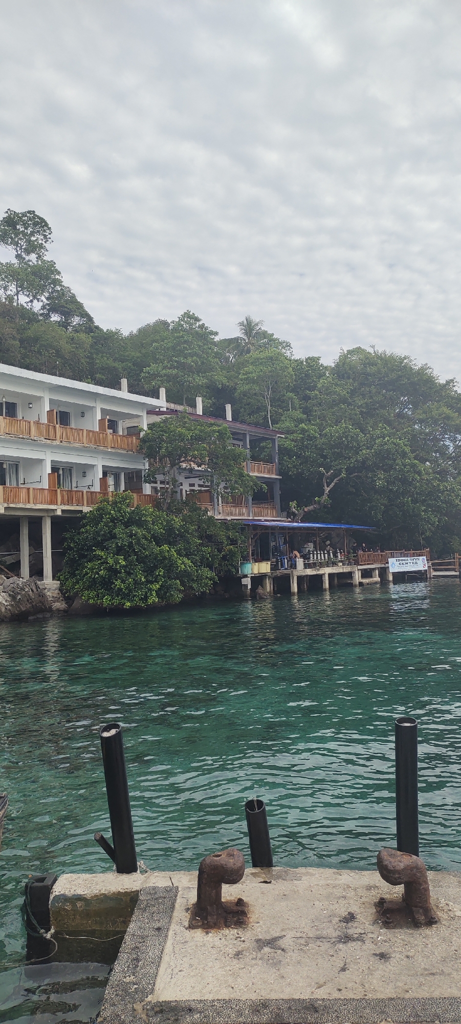 An outdoor, medium-shot, vertical-format photograph captures a serene waterfront scene in Sabang, Indonesia, with a building nestled on a tree-covered hill overlooking clear turquoise waters. In the foreground, a concrete pier juts into the water, equipped with two rusted bollards and two black pipes. The water is calm, with gentle ripples reflecting the overcast sky.
The middle ground features a multi-story white building with wooden balconies and railings, partially obscured by lush green trees. A small dock extends from the building, supporting a covered area with blue tarpaulin, where several people are visible. A sign on the right side of the dock reads "IBUH DIVE CENTER".
The background is dominated by a dense canopy of green trees covering a steep hillside, reaching towards a cloudy sky. The overall atmosphere is tranquil and suggestive of a tropical resort or diving center.