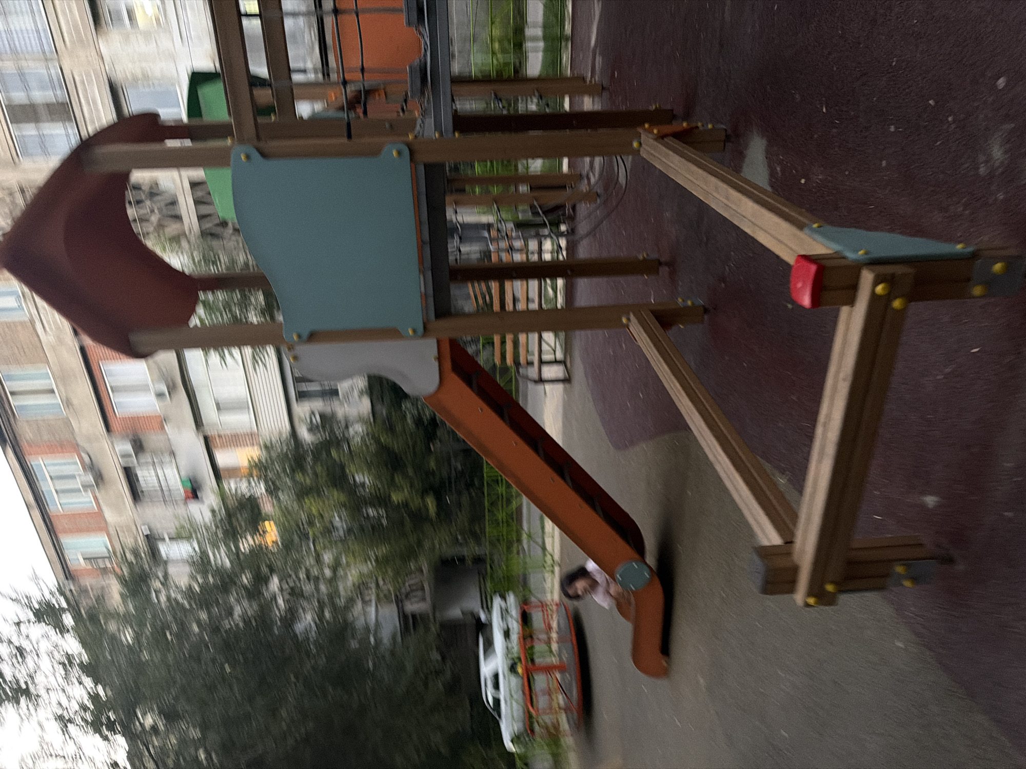 A young girl in a pink shirt is seated at the bottom of an orange slide on a playground in Shymkent, Kazakhstan. The playground features wooden structures and a red and orange merry-go-round with a white car on top in the background. An apartment building is visible behind the playground. The image is blurred, suggesting movement.