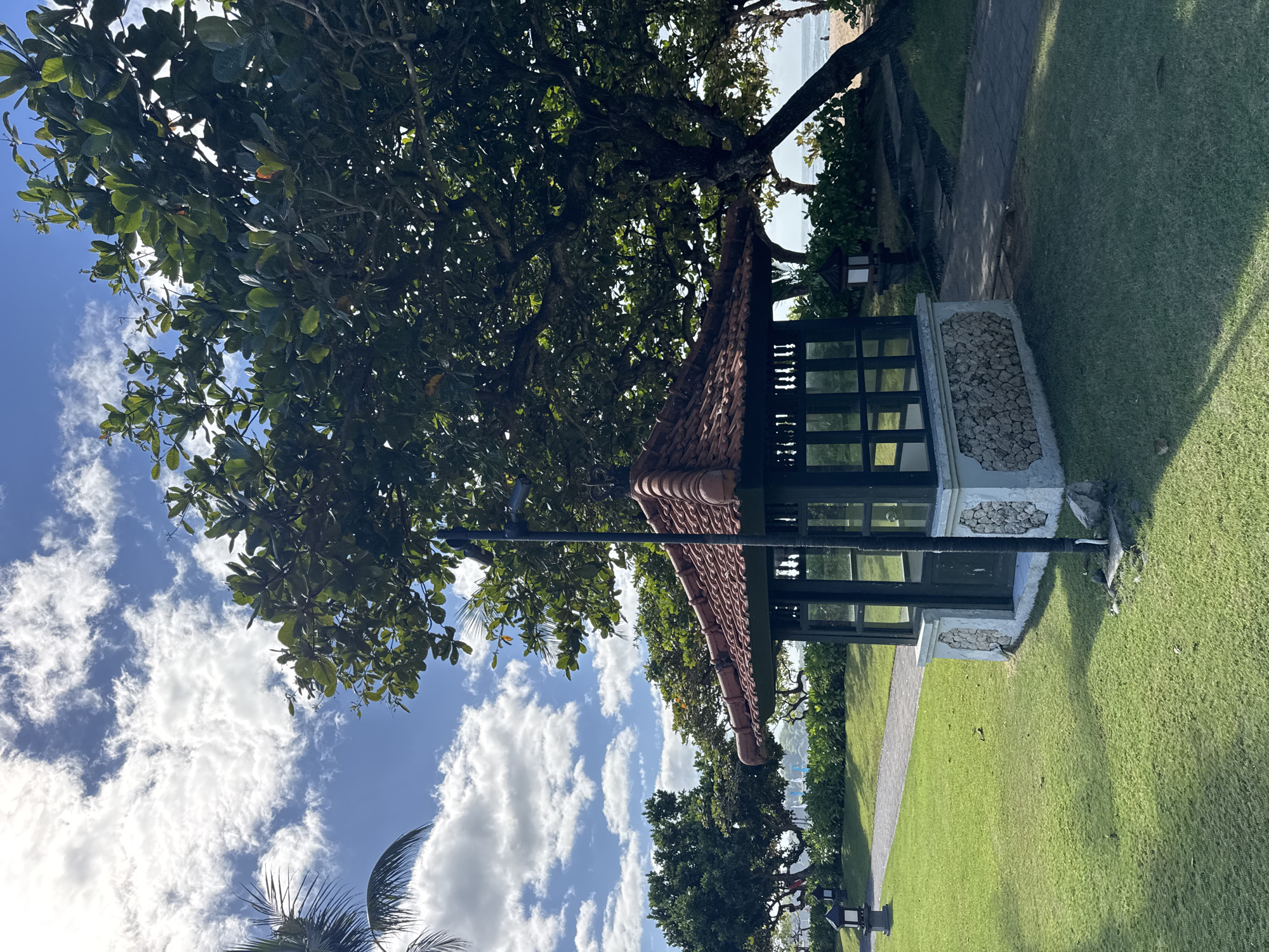 The image depicts a covered structure with a tile roof situated in a grassy area. The structure has black framing and some stone detailing on its base. It is partially obscured by a large tree with green leaves. To the right of the structure, there is a paved walkway adjacent to the grass. In the background, glimpses of a beach and the ocean can be seen. Above, a blue sky is partially covered with white, puffy clouds. The scene is likely located in Kuta Selatan, Indonesia, based on the tropical environment and architectural style.