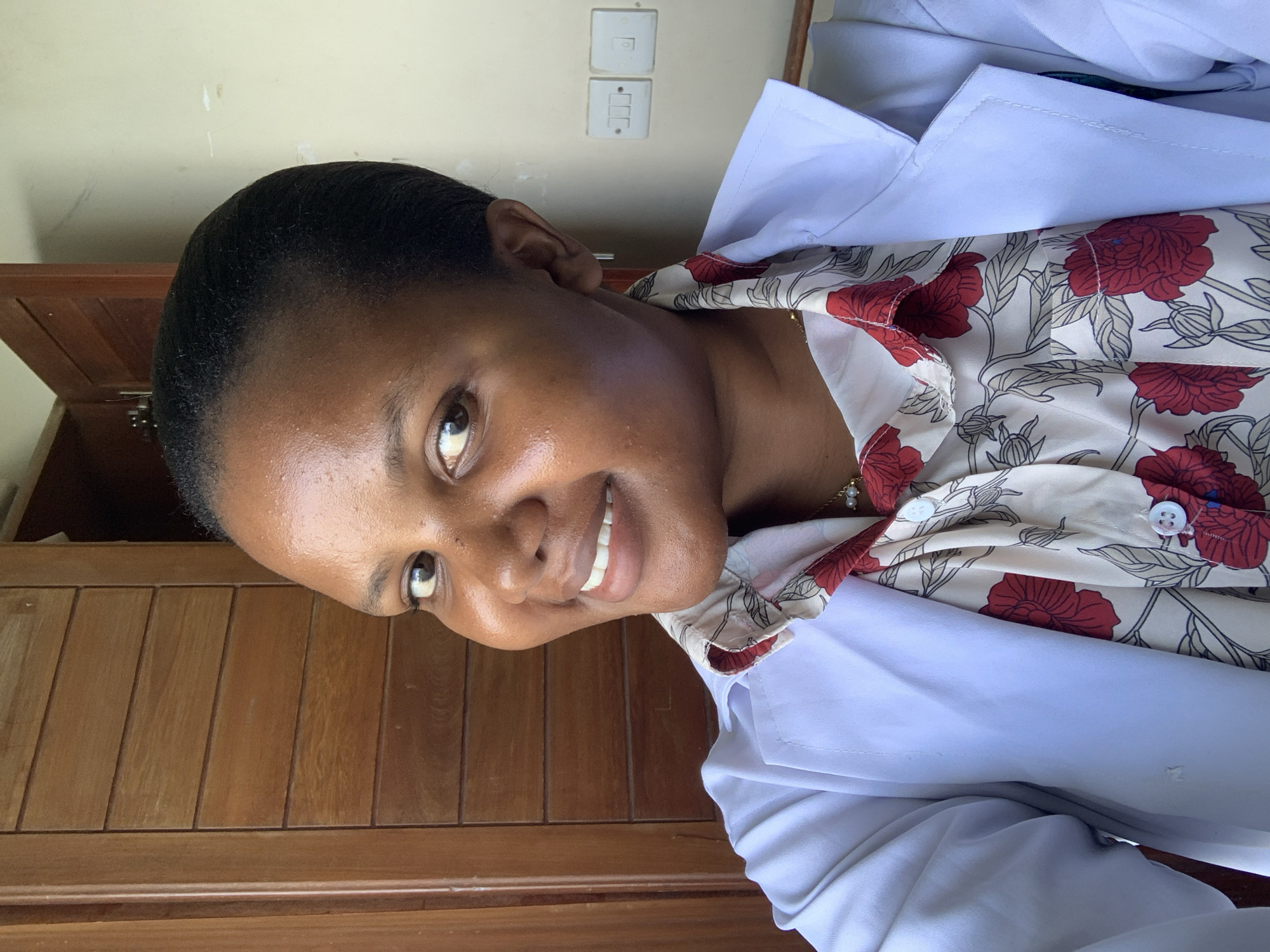 A person with dark skin and short black hair is smiling at the camera. They are wearing a white lab coat over a cream-colored shirt with red floral patterns. The background includes a light-yellow wall with electrical switches and a wooden cabinet. There is no information to confirm the location in Dar es-Salaam, Tanzania.