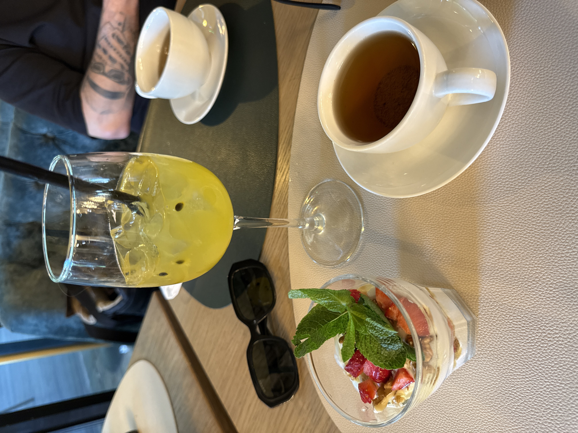 This image captures an overhead view of a table in what appears to be an indoor setting, likely a cafe or restaurant in Moscow, Russia. The scene depicts a casual meal or refreshment break with multiple drinks and a dessert.
On the left side of the frame, a person's tattooed arm, featuring a sleeve tattoo and a band tattoo, is partially visible. Next to the arm is a white teacup with a saucer, containing a light-colored beverage. This area rests on a dark green textured placemat. A tall glass with a vibrant yellow, ice-filled drink, possibly passion fruit juice given the visible dark seeds, is prominently featured with a black straw.
To the right, a white teacup and saucer hold a dark brown beverage, likely tea, placed on a light beige textured placemat. Adjacent to this, a small glass contains a layered dessert or breakfast item, composed of white creamy layers, chopped red strawberries, walnuts, and garnished with fresh mint leaves. A pair of black sunglasses rests on a light brown wooden strip that separates the two distinct placemat surfaces. A clear wine glass stem and base are also visible on the light beige surface.
The lighting is even, typical of an indoor environment. No text is visible in the image. The overall impression is one of a relaxed moment, enjoying a selection of beverages and a sweet treat.