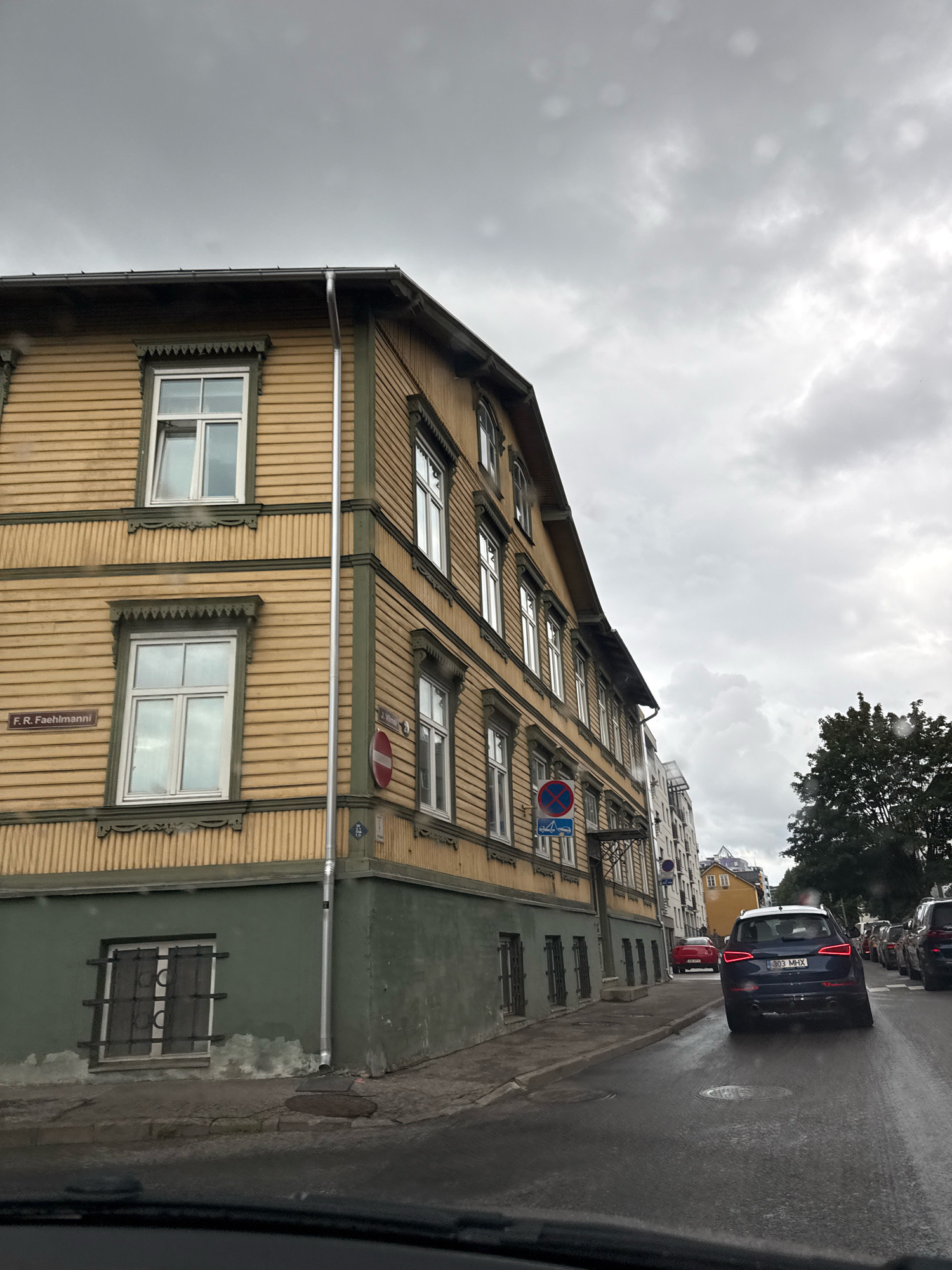 The image depicts a street scene in Tallinn, Estonia, likely in a historic district. The focal point is a yellow wooden building with green trim, characteristic of traditional Estonian architecture. The building has multiple windows, some with decorative elements above them. A street sign for "F. R. Faehlmanni" is visible on the building. The street is wet, suggesting recent rain. Several cars are parked along the side of the road or driving away from the viewer. One car in the foreground is a dark blue SUV, its license plate reads "303 MHX". The sky is overcast with heavy clouds. The overall atmosphere is subdued due to the weather.