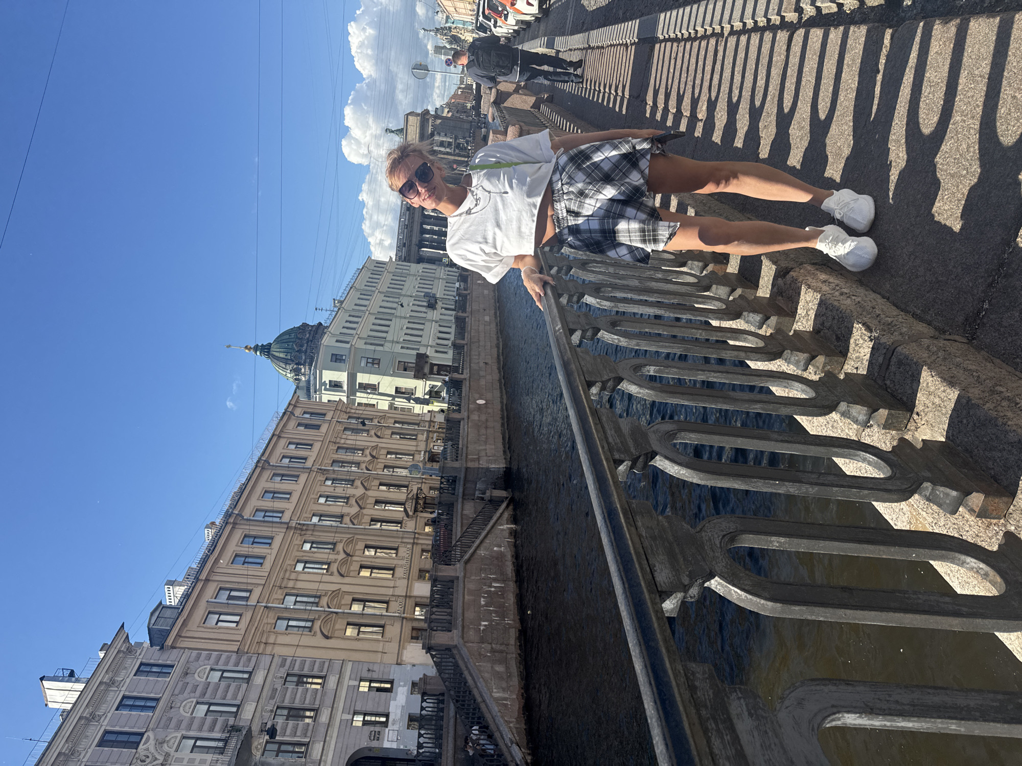 A woman stands on a bridge in Saint Petersburg, Russia, leaning on the railing and looking at the camera. She is wearing a white crop top, a plaid skirt, and white sneakers. Behind her, a canal runs through the city, with ornate buildings lining its banks. The sky is blue with scattered clouds, and the sun is casting shadows on the pavement. A man in a backpack is visible walking in the background on the right side of the image. The bridge features decorative ironwork and is made of stone. The architecture of the buildings suggests a historic European city.