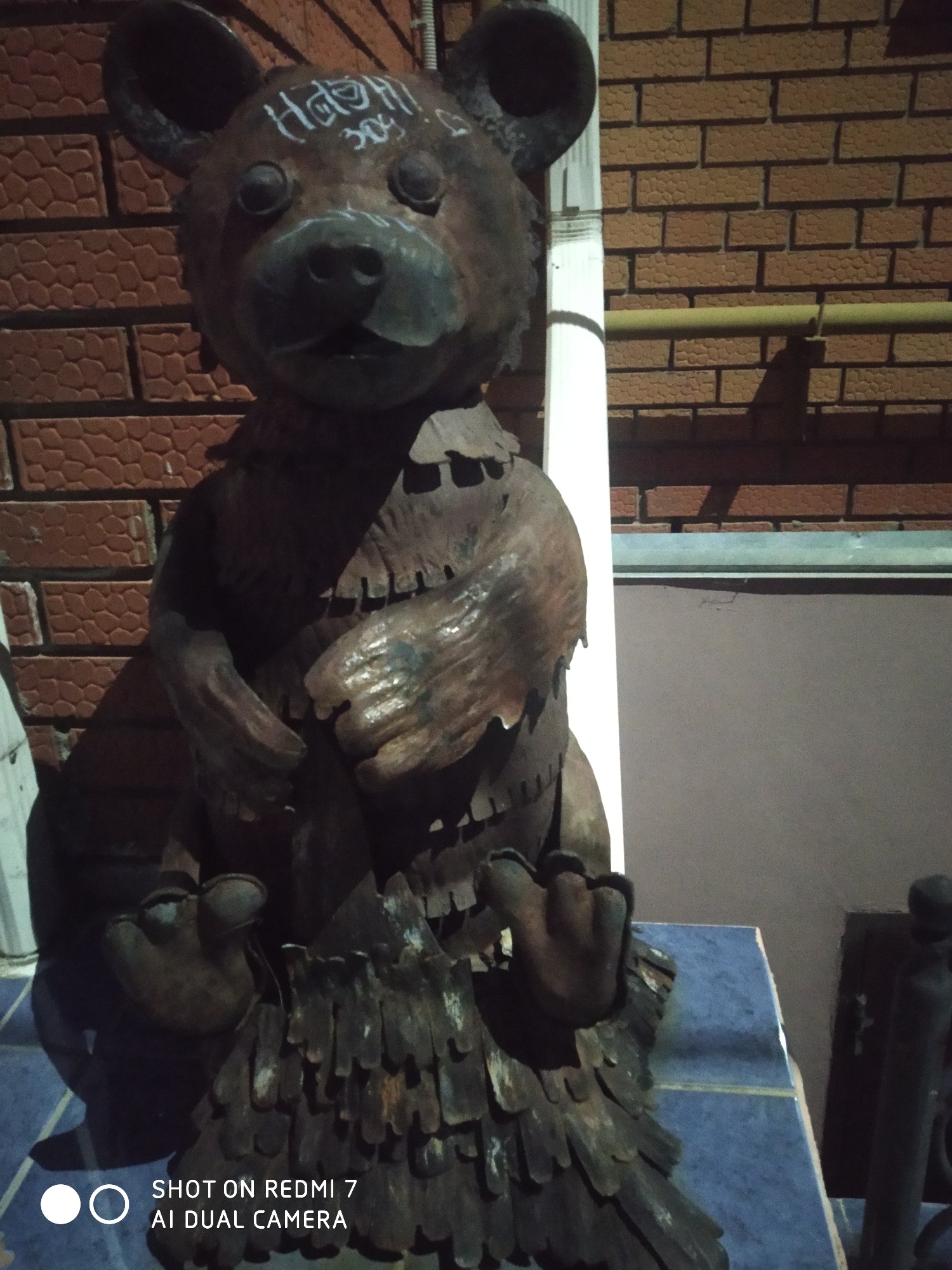 A metal bear statue with graffiti is situated in an outdoor environment. The statue is positioned against a brick wall, with a white pole running vertically adjacent to it. The statue has the word "HAHS!!" and the numbers "304" written on its head. The statue has a rough, fragmented texture, especially around its lower torso, which appears like layered metal pieces. The statue is sitting on a tiled surface. The photo was taken on a Redmi 7 AI Dual Camera. The location is Chaikovsky, Russia.