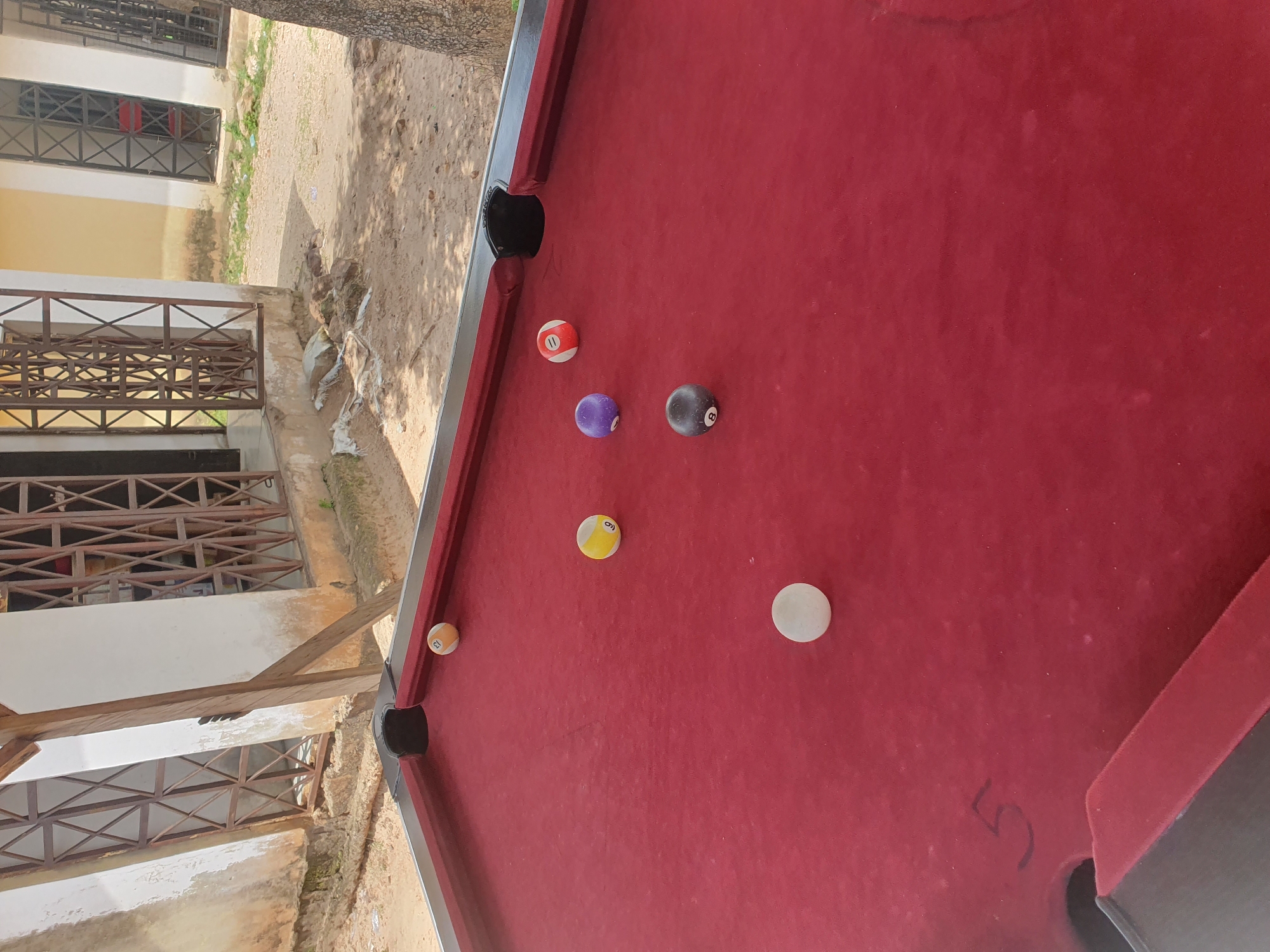 The image shows a close-up, angled view of a red billiard table with several pool balls scattered on its surface. The scene is outdoors, with a sandy ground and parts of a building visible in the background. The building appears to have multiple stories with barred windows and a pale yellow facade. Wooden structures and metal railings are also visible, suggesting an unfinished or rustic construction.
There are seven pool balls visible:
- A ball with the number "11" and red and white stripes.
- A solid purple ball.
- A solid black ball with the number "8".
- A solid white ball.
- A yellow ball with the number "6" in black.
- A yellow and orange striped ball with the number "13".
- A solid orange ball with the number "12".
One of the corner pockets of the billiard table is visible, and a number "5" is faintly visible, possibly drawn on the felt. The lighting suggests it is daytime, and the overall setting appears to be a casual outdoor area, potentially a yard or a patio. The presence of a billiard table outdoors in a location like Lokoja, Nigeria, suggests a recreational area. No people are visible in the image, and there are no clear indicators of the weather.