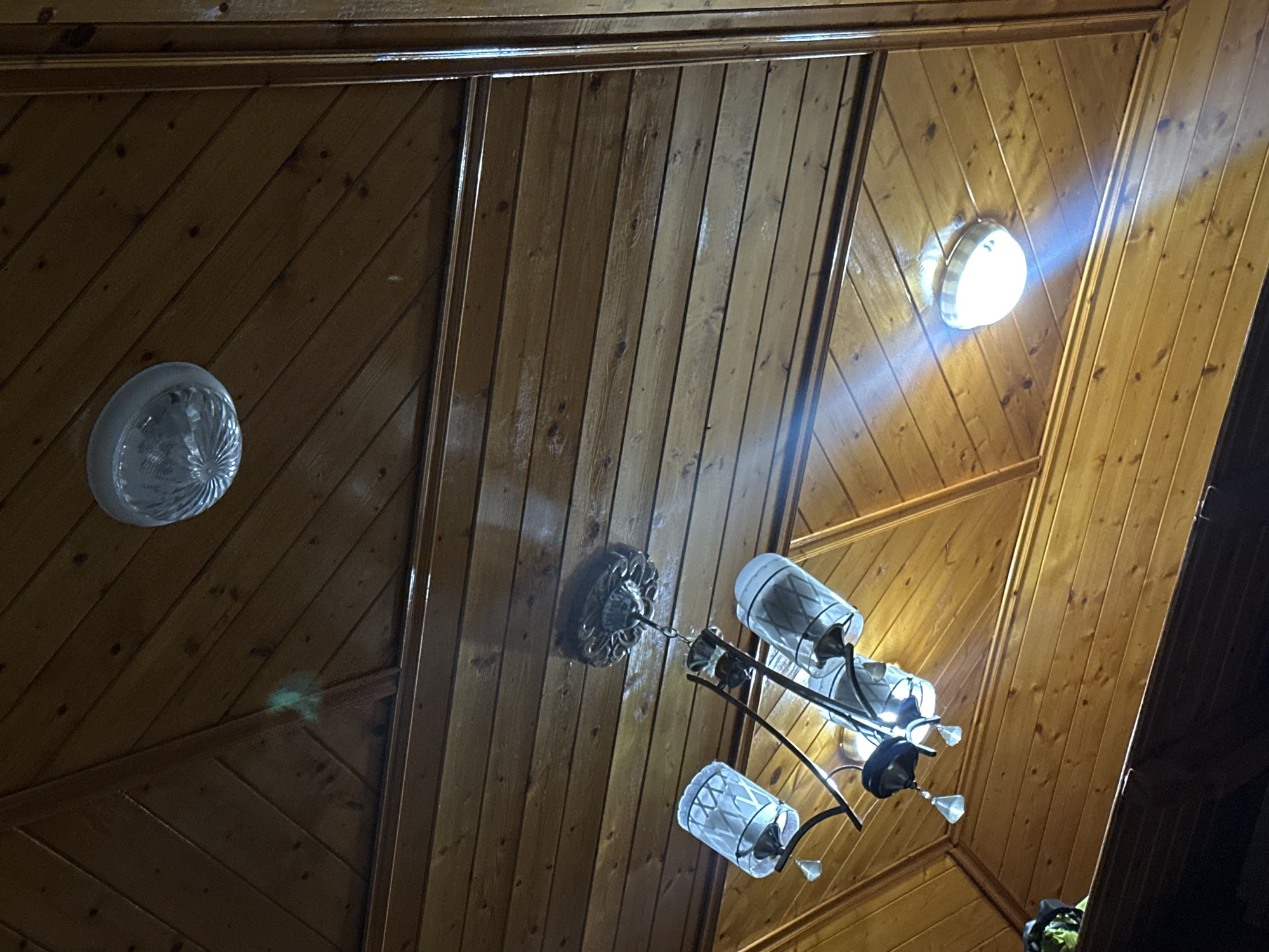 The image shows an interior shot of a wooden paneled ceiling, likely in the city of Berat, Albania. The ceiling is constructed from angled wooden planks and features two light fixtures. One is a semi-flush mount ceiling light with a clear, faceted diffuser, located in the upper right portion of the frame. Below and to the left of this is a decorative chandelier with four glass shades and metal accents, hanging from a central medallion. Another simple, circular light fixture is visible in the lower right quadrant. A faint green anomaly, possibly dust or a lens flare, is present on the wooden planks in the upper left. The overall impression is one of a rustic or traditional interior design.