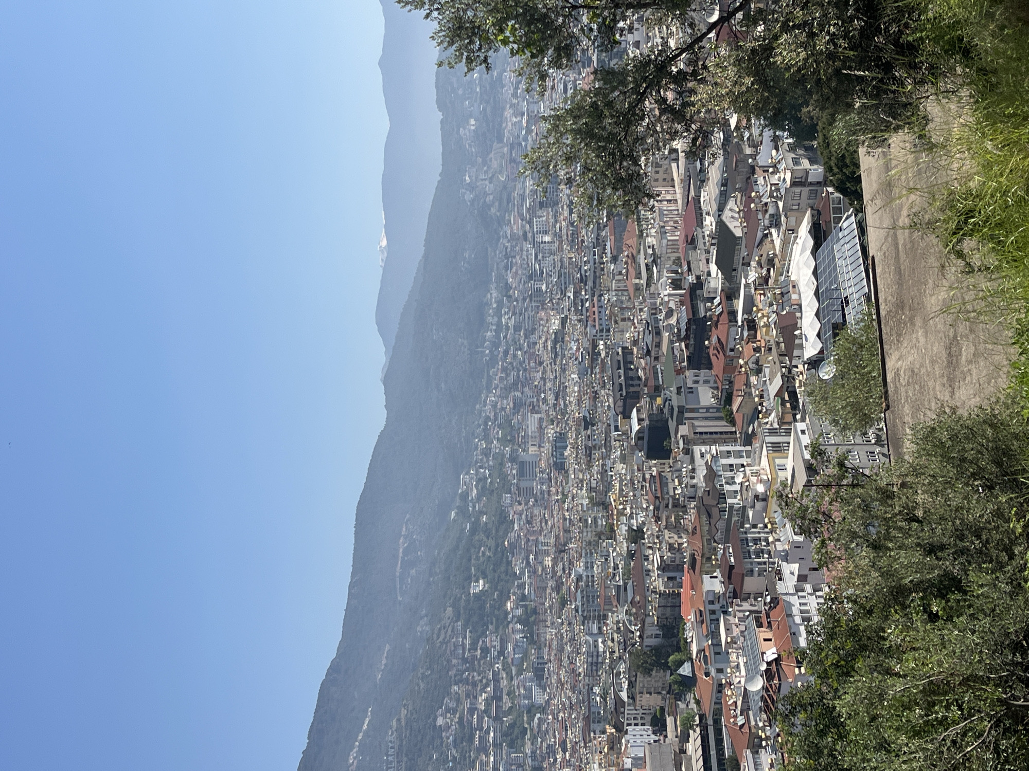 This image captures an expansive, high-angle view of the city of Alanya, Turkey, sprawling across a wide plain and extending up the lower slopes of a significant mountain range. The cityscape in the midground is densely packed with countless buildings, showcasing a mix of architectural styles. Many structures feature characteristic red-tiled roofs, while others appear more modern with flat roofs and some reflective glass facades.
The background is dominated by successive layers of mountains, which recede into a gentle atmospheric haze. A distinct lighter patch, possibly a snow-capped peak or light rock formation, is visible on one of the distant mountain summits, adding depth to the panorama. The foreground on the right side of the frame is adorned with vibrant green foliage and a section of a concrete path or wall, indicating the elevated viewpoint from which the photograph was taken.
The scene is bathed in bright, clear sunlight, suggesting it was captured during the daytime, likely late morning or early afternoon. The sky is a serene, clear blue. No specific human activity or event is depicted, making it a static landscape and cityscape photograph. A notable detail, visible on one of the mountain slopes, is the faintly legible white text "ALANYA," serving as a clear geographical identifier. The image conveys the impression of a vibrant coastal city nestled against a dramatic mountainous backdrop.