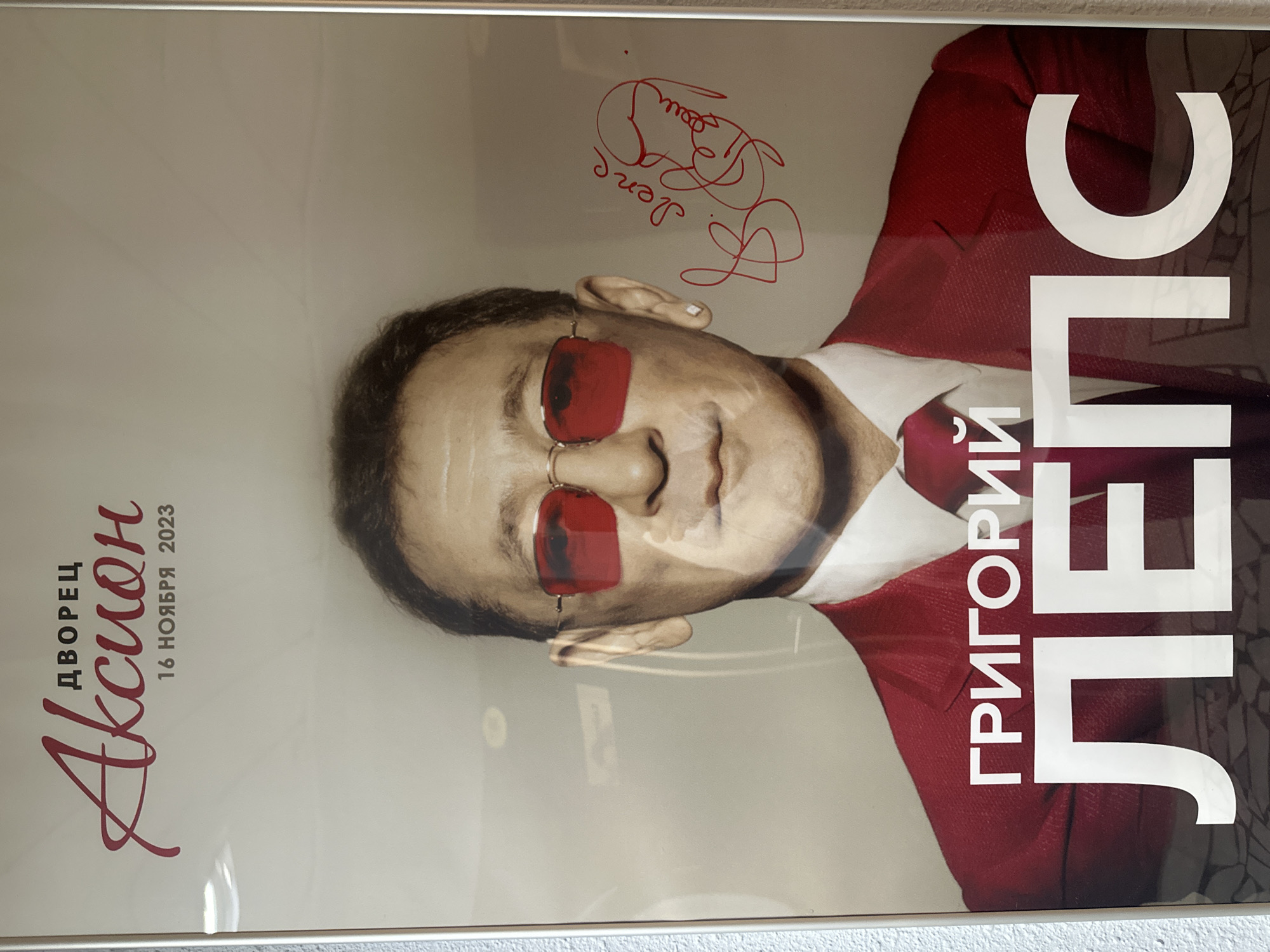 The image shows a framed poster for a Grigory Leps concert, held at the "Aksion" Palace in Izhevsk, Russia, on November 16, 2023. The poster features an image of Grigory Leps wearing red sunglasses and a red suit with a white shirt and red tie. The text on the poster includes "ДВОРЕЦ Акцион" (Aksion Palace), "16 НОЯБРЯ 2023" (November 16, 2023), and "ГРИГОРИЙ ЛЕПС" (Grigory Leps). The poster also has a red signature that reads: "Деня Лёша".