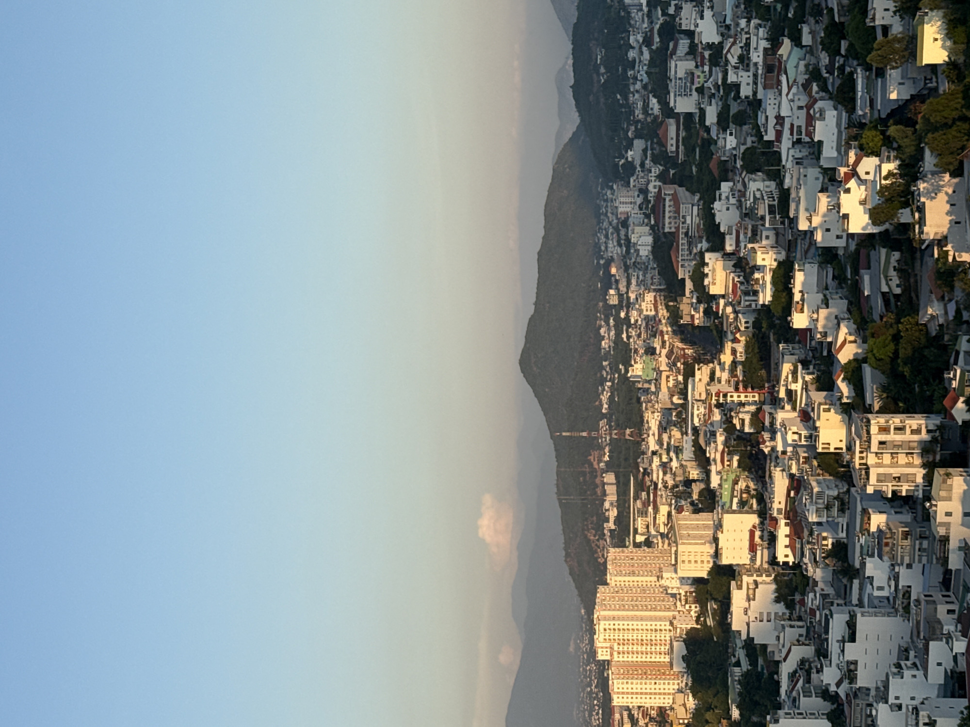 This is an aerial, landscape photograph of a sprawling city with rolling, green hills in the background. The sky is clear and a pale blue with a gradient towards a lighter hue near the horizon. The sun is low, casting a warm, golden light across the city, highlighting the buildings and creating long shadows.
In the foreground, a dense collection of buildings fills the frame. These structures vary in height and architectural style, with many appearing to be residential apartments and houses. Prominent in the left mid-ground is a tall, cream-colored apartment building with numerous balconies. Adjacent to it are shorter, white buildings with red-tiled roofs and flat-roofed structures of various sizes. The buildings are tightly packed, with narrow streets and alleys visible between them. Lush green trees are interspersed throughout the urban landscape, particularly in the lower portions of the image.
In the mid-ground, more buildings are visible, some appearing to be commercial or administrative, with a prominent red and white radio tower rising above them. Behind the city, a range of green, tree-covered hills form a natural backdrop. One of these hills has a distinct, rounded peak. A few wispy clouds are present in the sky above the hills. The overall impression is a city nestled within a verdant, hilly terrain, bathed in the soft light of either sunrise or sunset. The scene appears to be a typical urban view of Nha Trang, Vietnam, characterized by its coastal location and surrounding natural beauty.