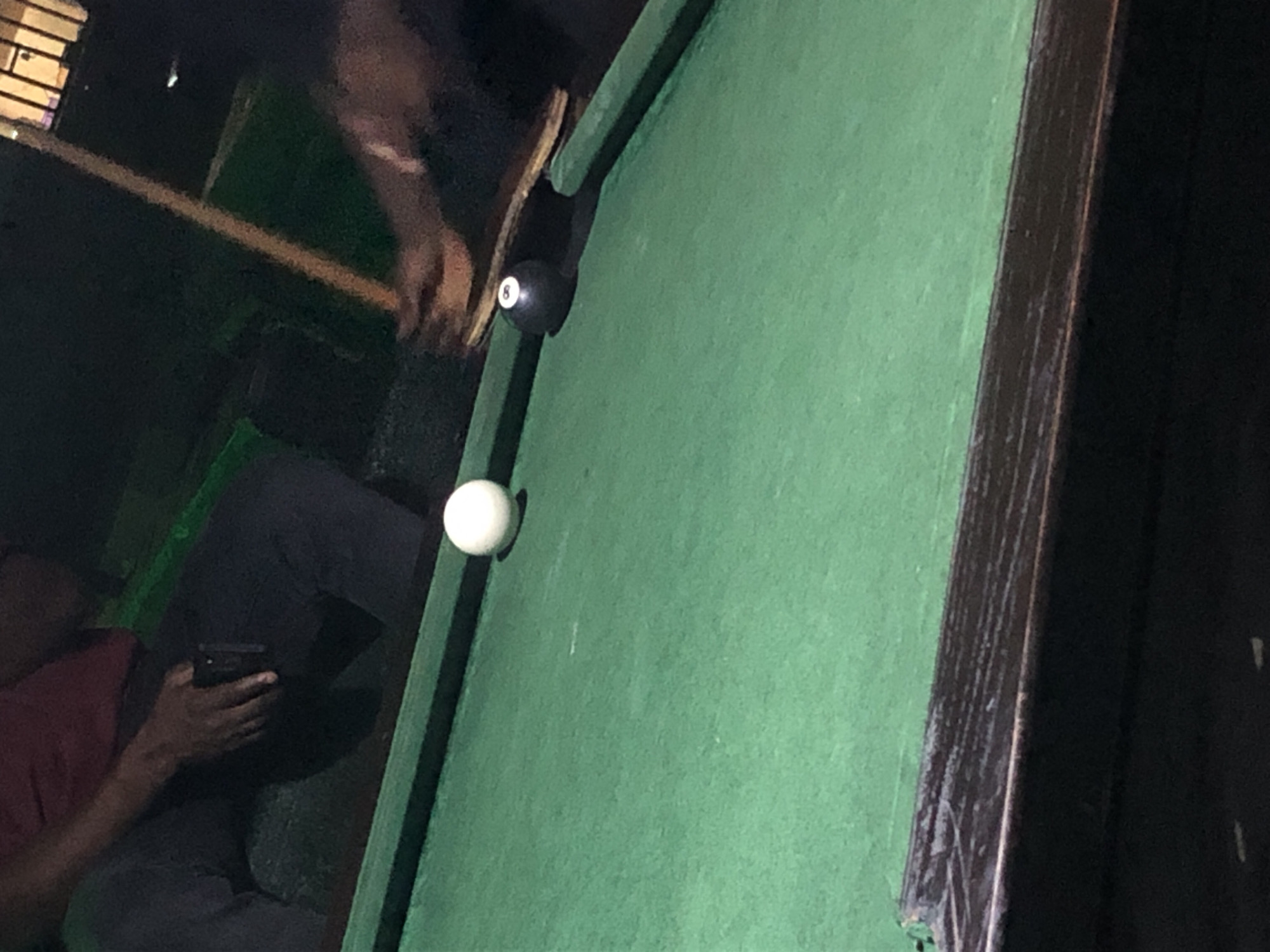 The image depicts a close-up view of a green billiard table with two balls. A white cue ball is positioned in the upper left quadrant of the table, while a black eight ball, marked with the number "8", is situated to its right. A pool cue stick rests diagonally across the table, near the eight ball, with hands visible on the cue, suggesting someone is playing. The background is dark and indistinct, with hints of furniture and possibly other people. The scene appears to be indoors, likely in a dimly lit establishment such as a bar or pool hall. The specific location in the city of Unknown, Unknown is not discernible from the visual information provided.