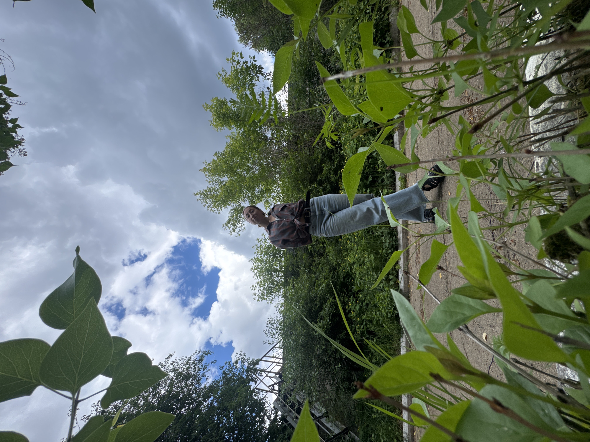 A vertically oriented photograph depicts a person standing on a concrete surface, surrounded by dense greenery, against a backdrop of a cloudy sky. The person is wearing a plaid shirt, jeans, and black shoes. They are positioned in the middle ground, facing the camera with a slight smile. The foreground is filled with large green leaves and stems, creating a sense of depth. The background includes trees, vegetation, and a partly cloudy sky. The photograph gives the impression of being taken at ground level, looking upwards. The location is in the city of Chaikovsky, Russia.
