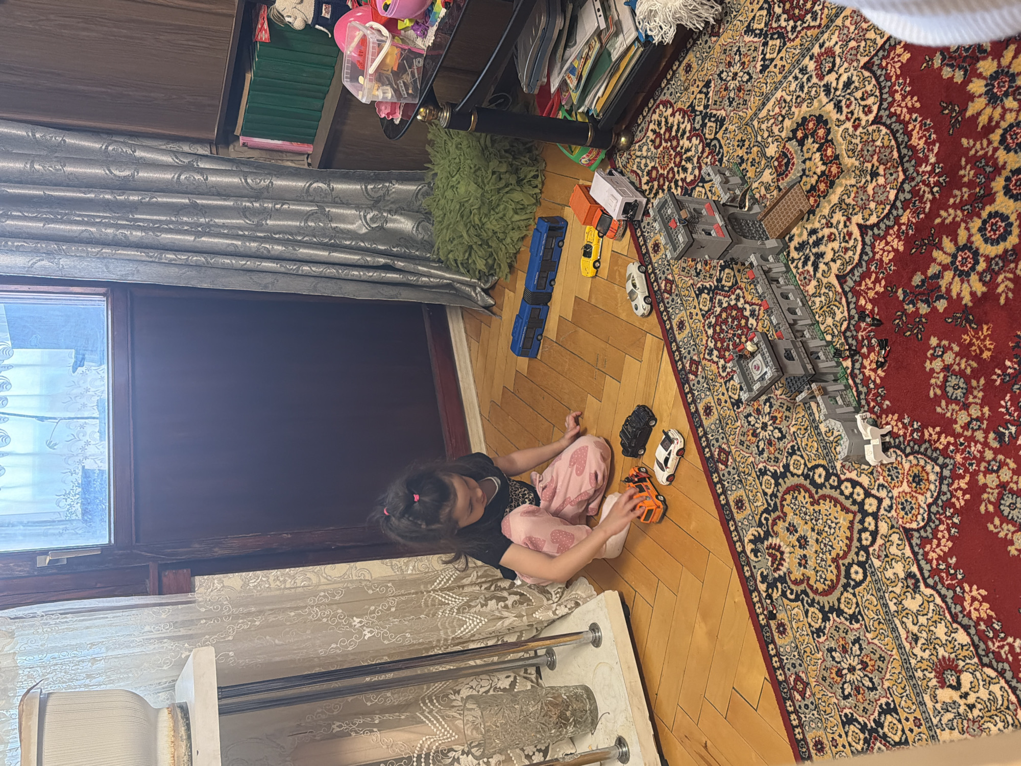 A young girl is seated on a wooden floor next to a white cabinet and playing with three toy cars. In the background, a red patterned rug covers the floor next to a partially built Lego castle. Beside the castle are additional toy vehicles, including a blue articulated bus, a yellow car, and an orange vehicle. Above the Lego construction, there is a glass-topped shelf unit filled with books, containers, and other items. A fluffy green rug is placed near the base of the shelf. To the left, there is a window covered by a silver curtain. The location is Марковский, Russia.