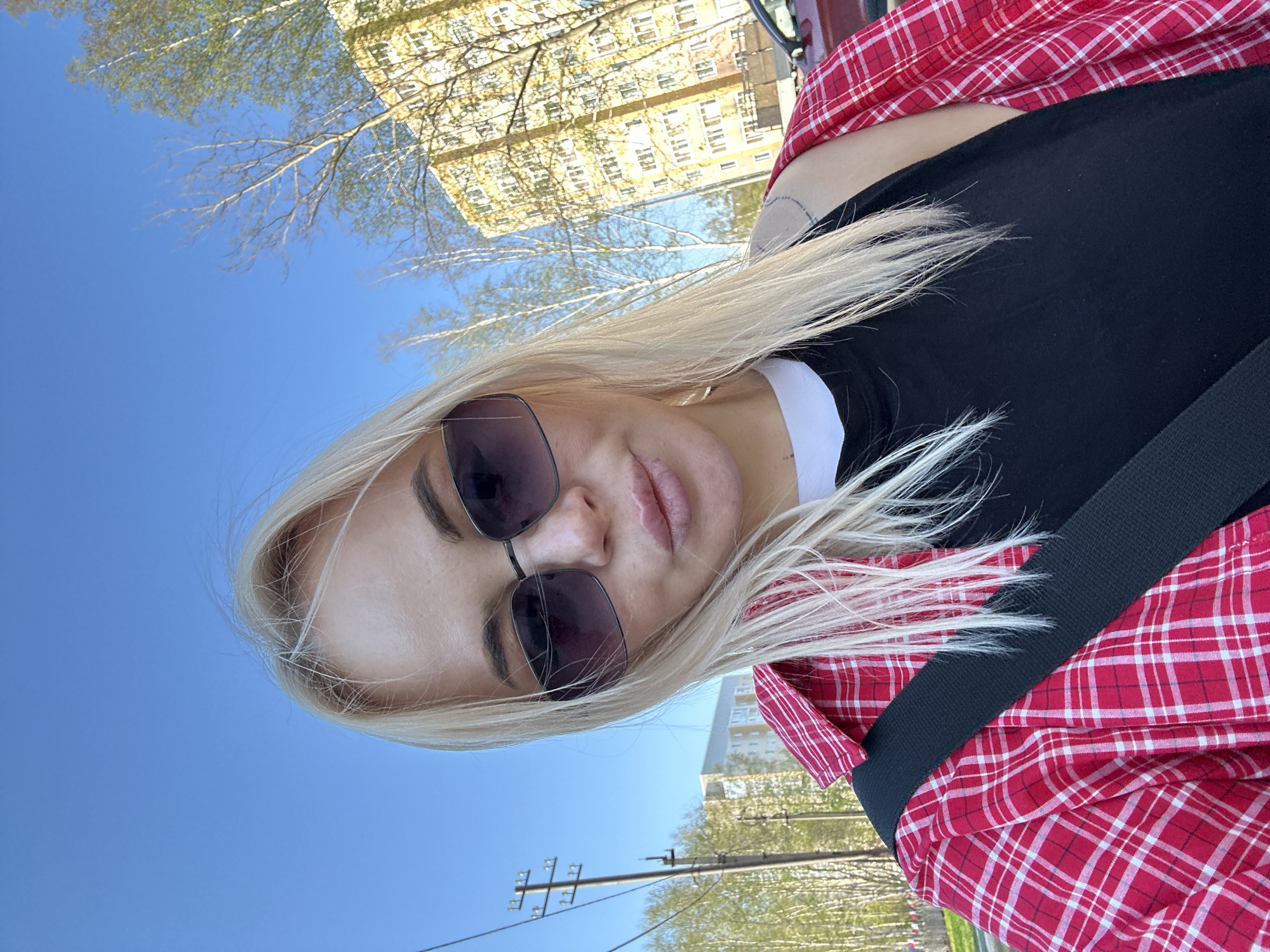 A woman with blonde hair and sunglasses is pictured from a close, upward angle. She is wearing a black shirt with a white collar and a red and white plaid shirt. A black strap, possibly a seatbelt, crosses her body. In the background, there is a clear blue sky, buildings, and trees with budding leaves. Electrical poles are also visible. The location is Megion, Russia, as identified by the surrounding environment.