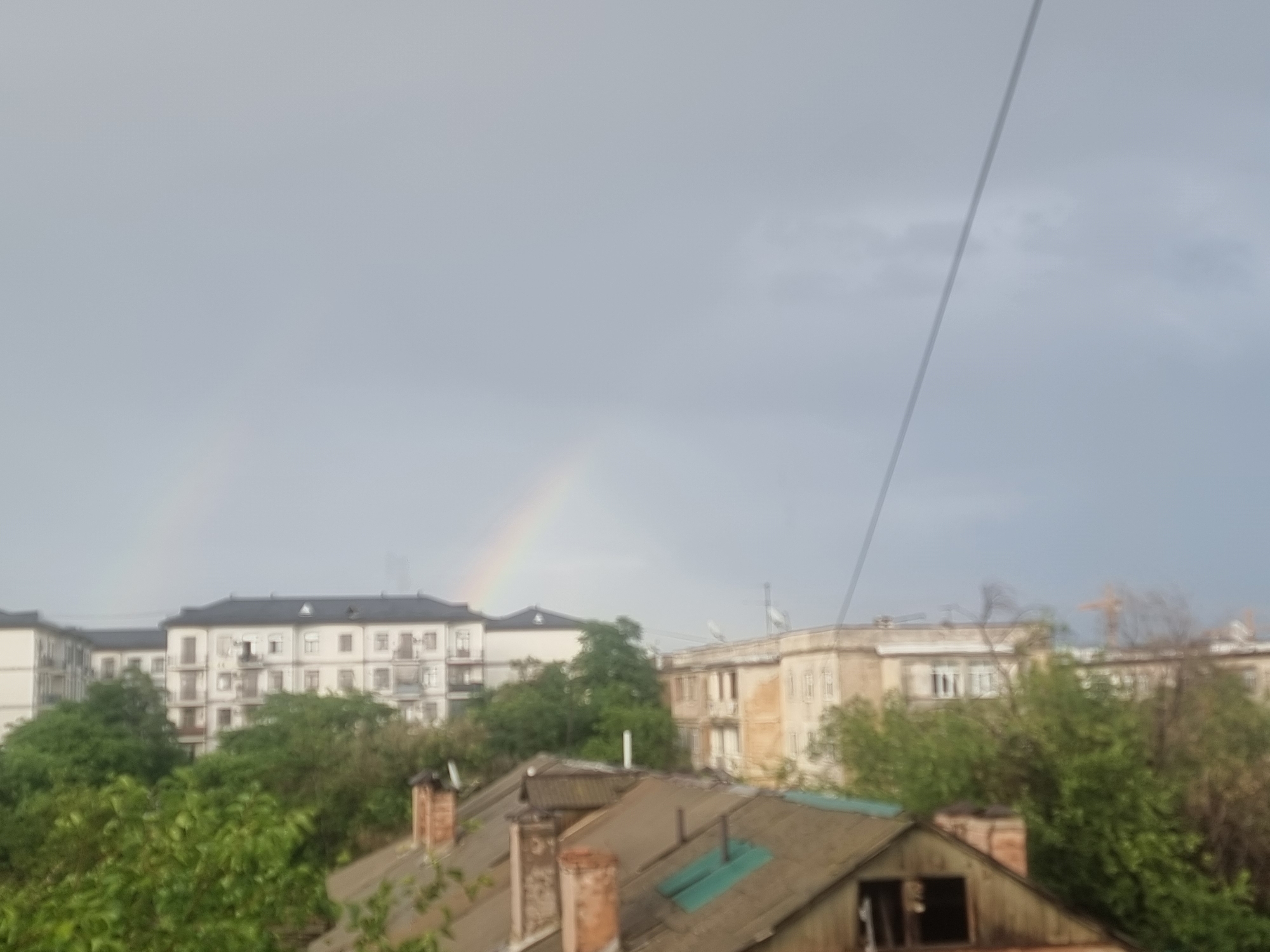 This media file depicts an overcast sky with a faint rainbow visible. In the foreground, the rooftops of several old buildings are visible, some with chimneys. Behind these rooftops, trees and apartment buildings can be seen. The apartment buildings appear to be in a residential area, with multiple windows and balconies. The overall atmosphere suggests a post-rain or stormy weather scene. The light suggests it is daytime, likely late afternoon or early evening given the muted light. The scene is likely set in Tashkent, Uzbekistan, as suggested by the architectural style of the apartment buildings and the presence of trees common in the region. No people are visible in the image.
