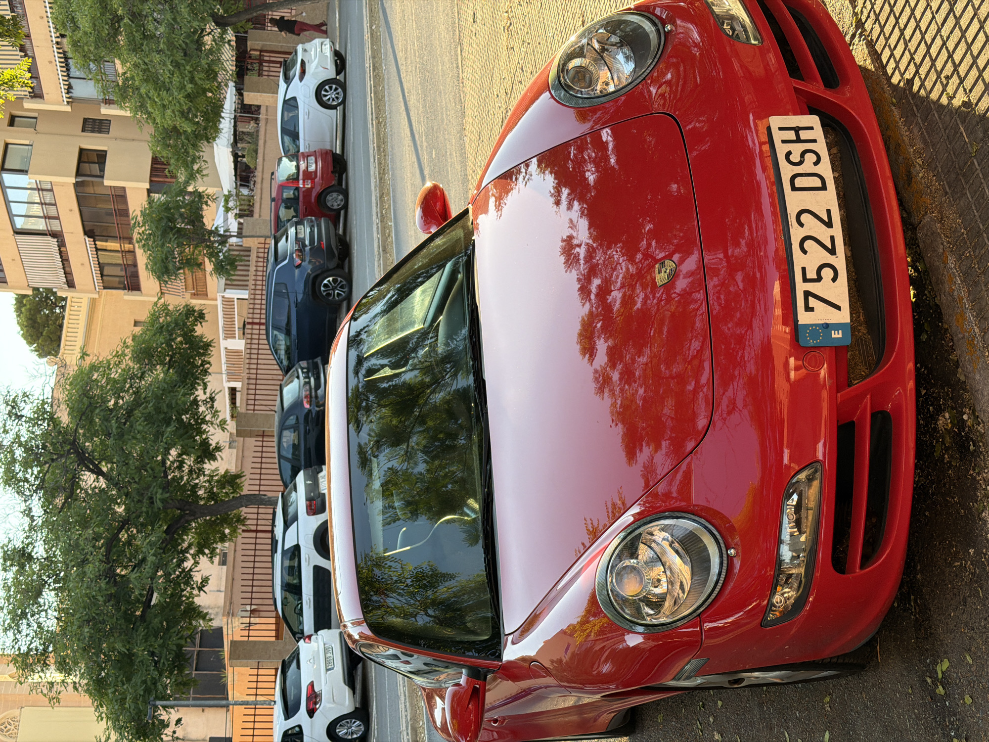The image shows a bright red Porsche 911 parked on a sun-drenched street. The car is in the foreground, with its front end prominently displayed. Its license plate reads "7522 DSH" and has the European Union emblem. The car's surface reflects the surrounding environment, including trees and buildings. In the background, several other cars are parked, including a white Citroen, a white Jeep, a small red car, and a white Opel. Buildings with balconies and awnings line the street, and a large tree with green foliage stands to the left of the frame. A person in red pants and a dark top is visible walking away from the parked cars on the right side of the image. The ground is paved with cobblestones in some areas and asphalt in others, with scattered fallen leaves. The overall scene suggests an urban setting, possibly a residential street.