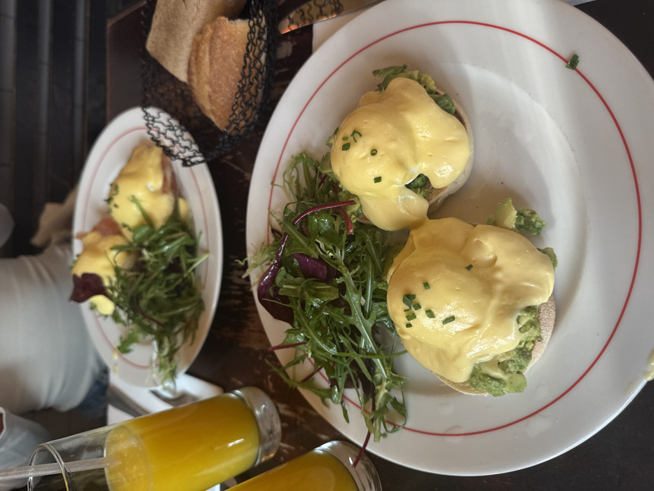 The image captures a dining table scene in a cafe or brasserie located in Paris, France. In the immediate foreground, a white plate with a red rim presents two portions of avocado Eggs Benedict, each consisting of an English muffin base, topped with mashed avocado, a poached egg, and hollandaise sauce, garnished with chopped chives. Adjacent to these is a fresh mixed green salad. To the left, a clear glass filled with orange juice contains a transparent straw.
Further back, a second white plate with a red rim holds two portions of traditional Eggs Benedict, appearing to feature ham beneath poached eggs, covered in hollandaise sauce and chives, also accompanied by a mixed green salad. To the right of this plate, a small black wire basket contains two pieces of light-colored bread, consistent with baguette portions. A metal knife with a dark, textured handle lies on the dark table surface beside the bread basket. A second glass of orange juice is partially visible on the far left. A person wearing a white top is indistinctly seated in the background, indicating a dining companion. The table surface is dark, likely wood or stone. The specific plating and meal presentation are characteristic of Parisian cafe dining.