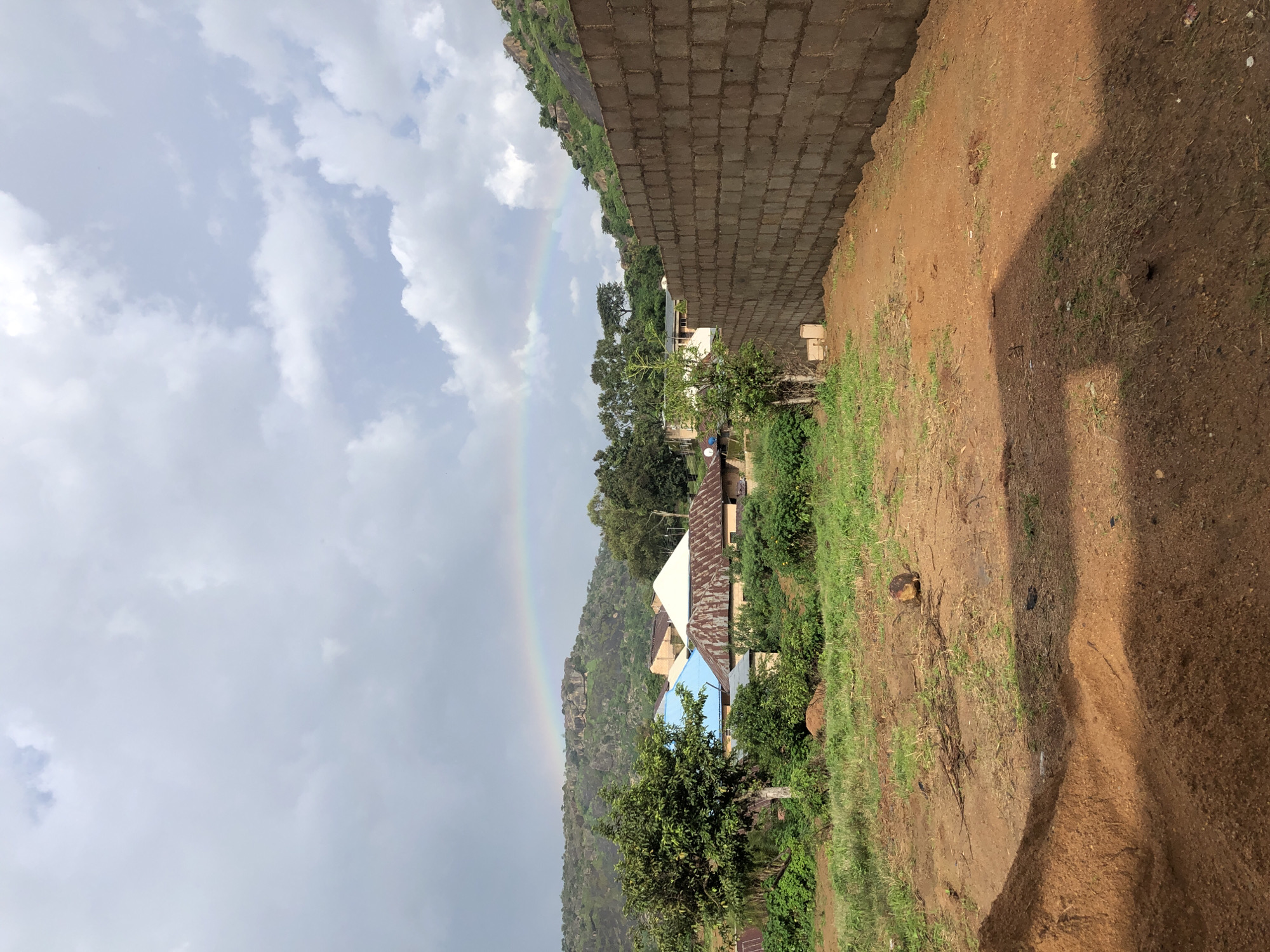 In Jos, Nigeria, a wide shot captures a rural landscape under a cloudy sky with a faint rainbow arching over a rocky, tree-covered hill. In the foreground, a pile of reddish-brown soil sits on unpaved ground, casting a shadow. To the right, a partially constructed wall of grey concrete blocks stretches across the scene. Beyond the foreground, green grass and scattered vegetation lead to a cluster of buildings with diverse roofing, including a blue-domed structure and a white-roofed house. Trees dot the middle ground, with a larger tree on the left. The background features the aforementioned rocky hill and more greenery. The overall impression is one of a developing area with natural beauty.