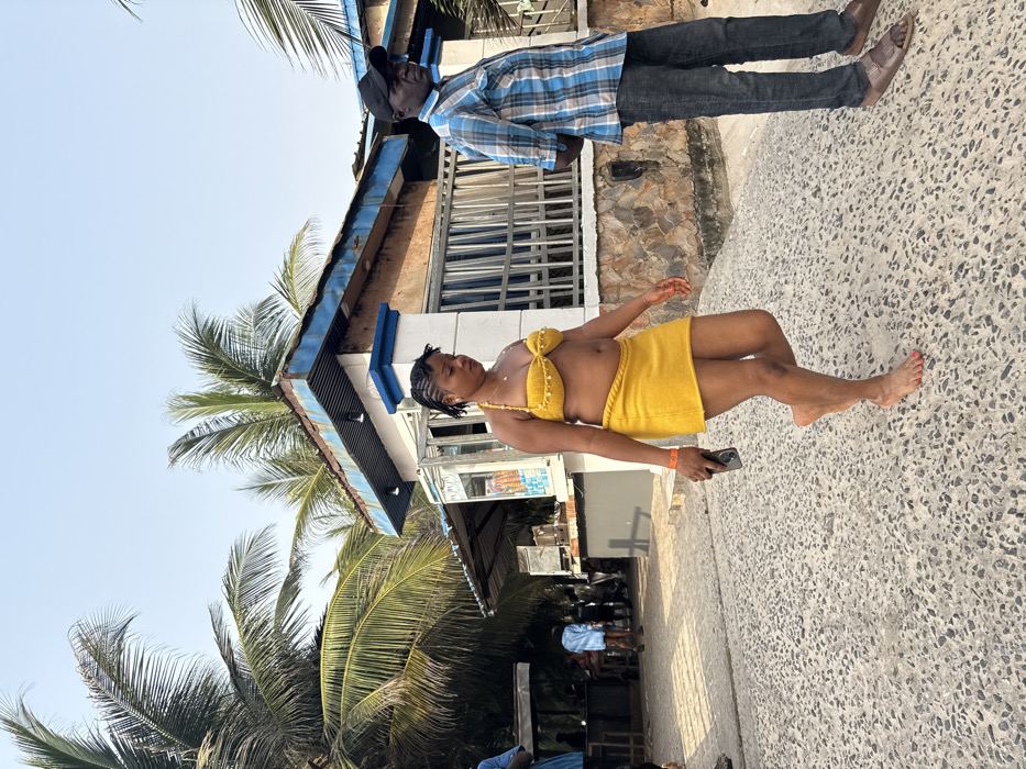 A woman in a yellow two-piece outfit stands on a concrete surface next to a building with palm trees. She is holding a cell phone in her left hand. Another person, wearing a plaid shirt and dark pants, stands next to the building. The building has a blue roof and stone accents.