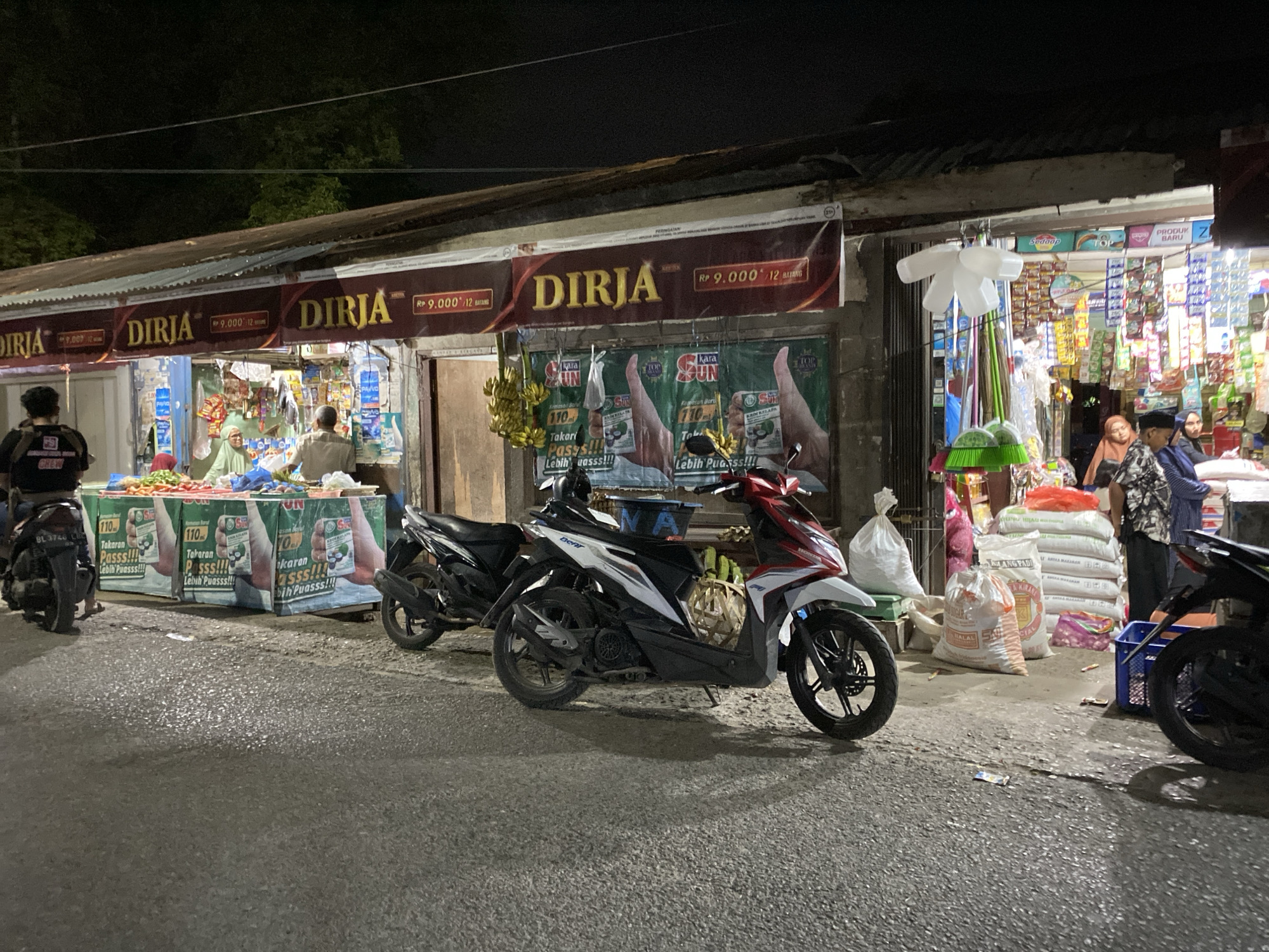 In an outdoor market scene at night, several motorcycles are parked along a street in front of a row of stalls. The stalls display various goods, including bags of rice and other items on shelves. A banner overhead reads "DIRJA" with pricing information. People are visible behind the stalls, interacting with customers or arranging merchandise. The ground is asphalt, appearing wet and reflecting the light from the stalls. The overall atmosphere suggests a busy, open-air marketplace operating after dark.