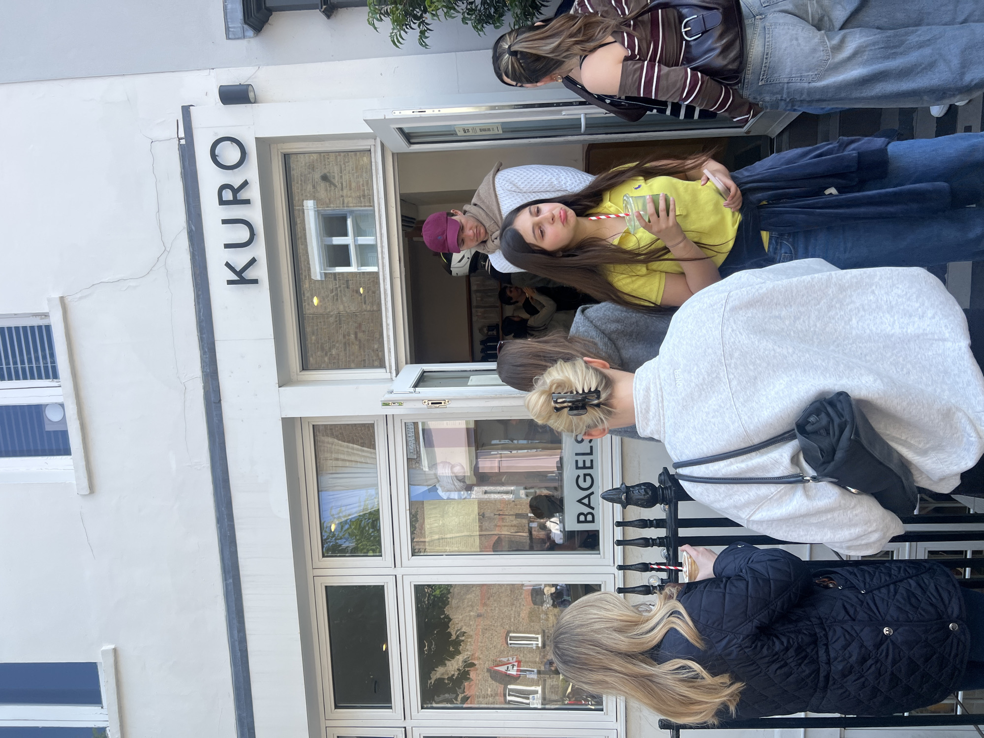 A vertical, medium-angle shot depicts a group of people standing in front of a building in London, United Kingdom. Above the open doorway, the name "KURO" is displayed in black letters. A sign for "BAGELS" is visible in the window of the building. The people are standing on the sidewalk in front of the building. Some individuals are holding drinks. The environment appears to be a bustling urban setting.