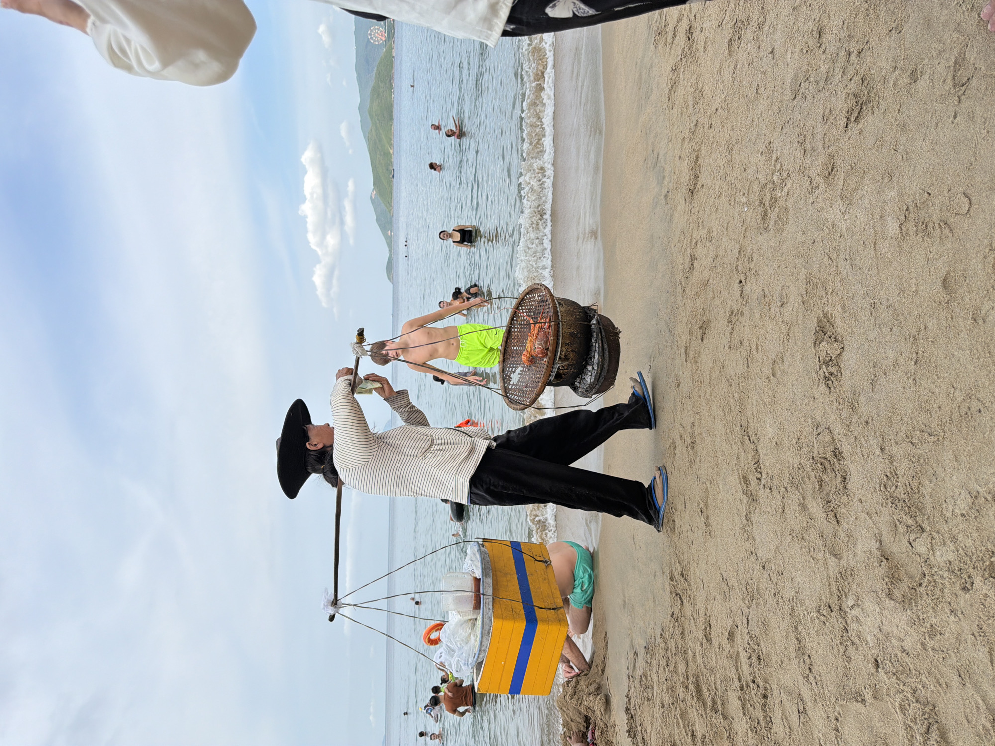 A person wearing a black conical hat and a striped shirt carries food on a yoke across a sandy beach in Nha Trang Ward, Vietnam. On the left side of the yoke, a yellow and blue container holds food items. On the right side, a basket with grilled seafood sits above a charcoal brazier. In the background, people are swimming in the ocean under a cloudy sky. The scene depicts a vendor selling food on a crowded beach.