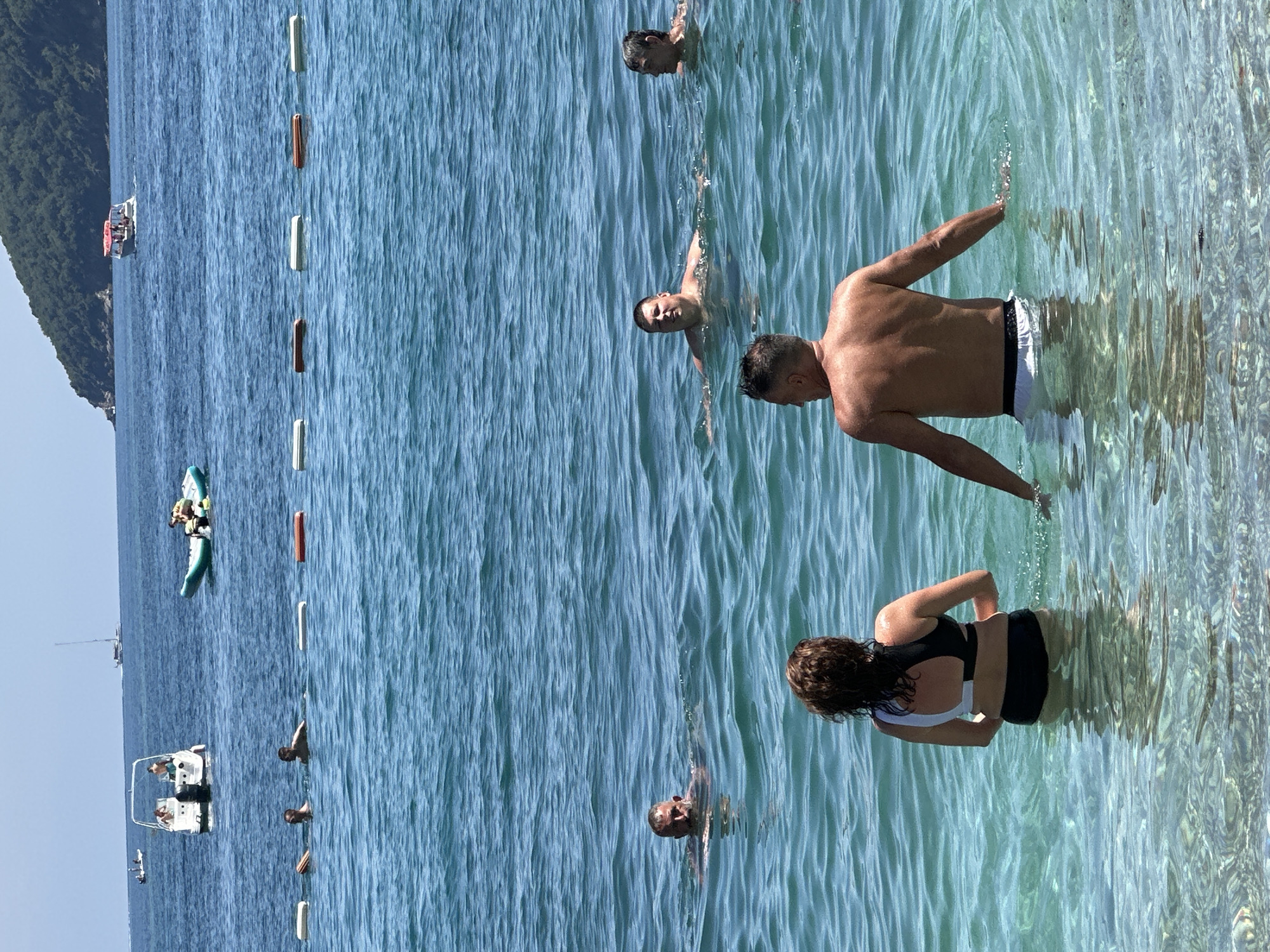 The image captures a sunny day at a beach in Budva, Montenegro. In the foreground, a man and a woman stand in clear, shallow water, facing away from the camera. The man is shirtless, wearing white swim trunks, and the woman is in a black and white bikini. Behind them, several men are swimming in the deeper, blue water. In the middle ground, buoys mark a designated swimming area. In the background, various boats are visible on the sea, including a speed boat with people onboard, a small boat with an outboard motor, a sailboat, and a banana boat with occupants. The coastline features a lush, green, mountainous landscape under a clear blue sky. The water is calm with small ripples.