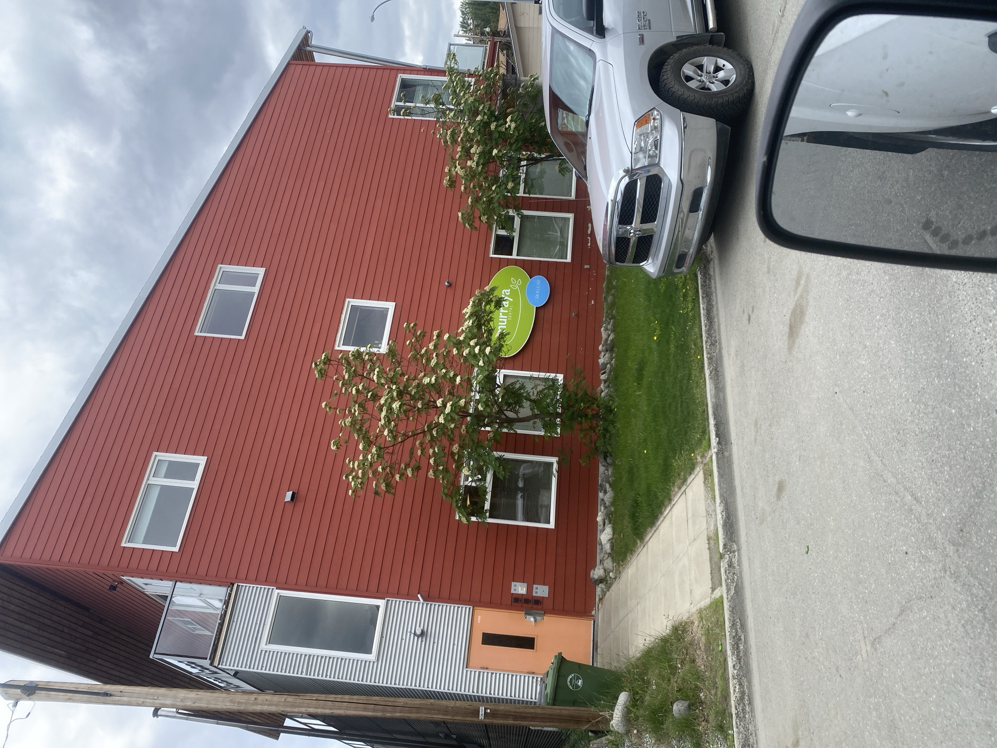 A silver Dodge Ram 1500 pickup truck is parked on a paved road in front of a two-story building with red siding and a sloped metal roof. The building has several windows and an orange door on the ground floor. A green trash bin is next to the door. There is a sidewalk and a grassy area between the building and the road. A green oval sign with white lettering is mounted on the building. The sky is overcast. The location is the Murray Dental clinic at 4969 4th Avenue in Whitehorse, Canada.