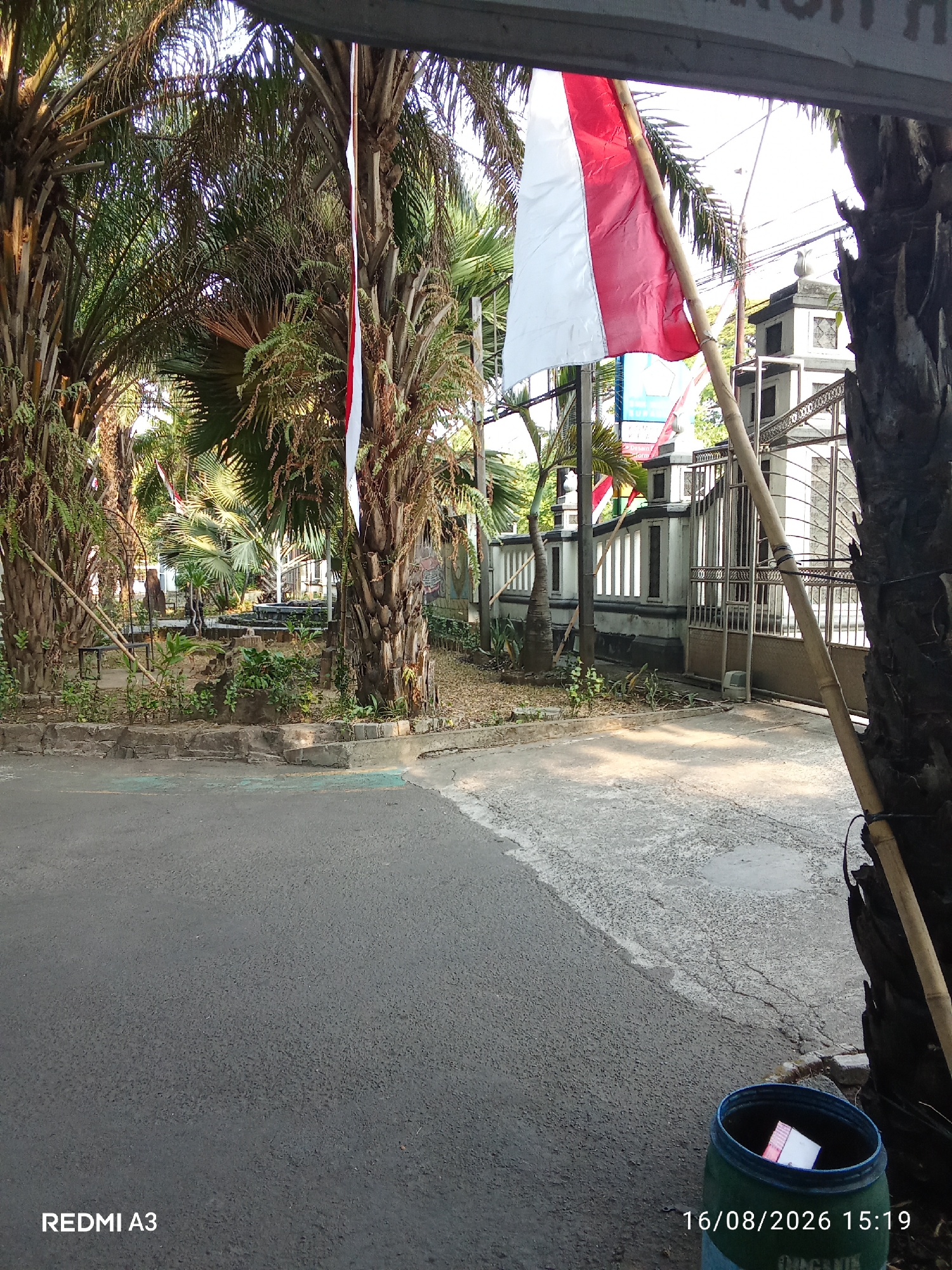 The image depicts an outdoor scene with palm trees in the foreground. A flag, likely the Indonesian flag, with red and white stripes, is visible, attached to a bamboo pole. Behind the trees, a white fence with metal bars and a gate can be seen, along with a building that appears to be a gatehouse or a small structure. The ground is paved with asphalt and concrete. There is a green trash can with some items inside, located on the right side of the image. The overall impression is that of an entrance to a property in a tropical or subtropical environment. The text overlay "REDMI A3" and "16/08/2026 15:19" indicates the device used for capture and the date and time. This specific view is likely located in Surakarta, Indonesia, given the presence of the Indonesian flag and the prevalent vegetation.