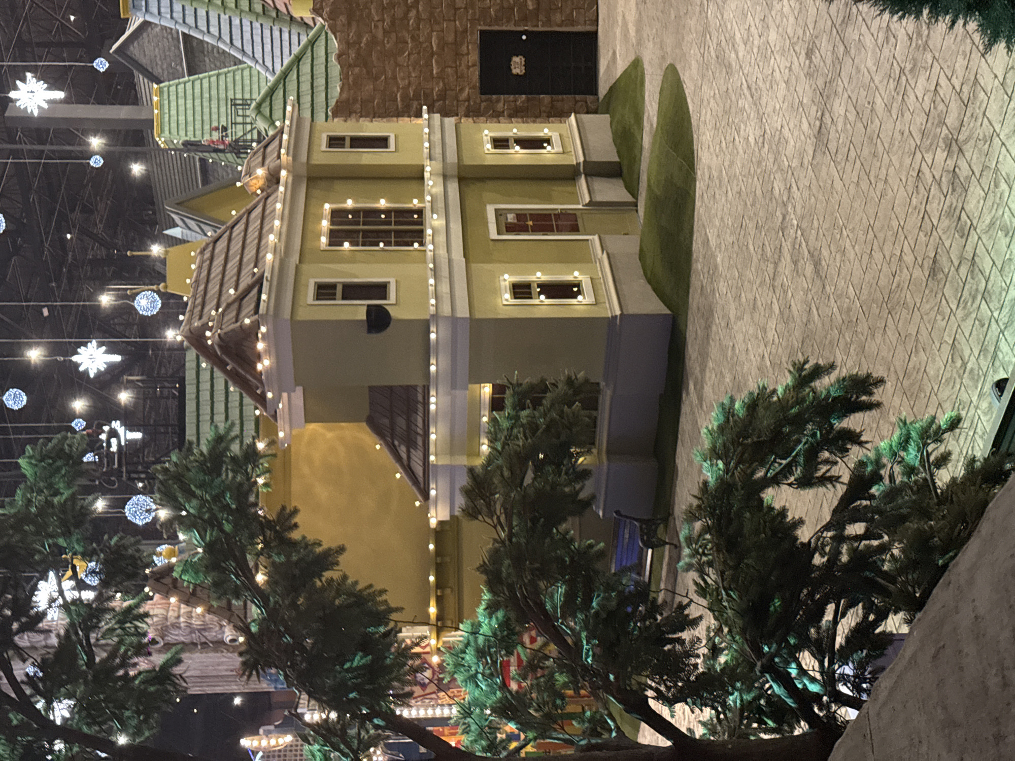 The image shows a building with a green and yellow exterior, decorated with Christmas lights. It has a brown roof and several windows. A black door is visible on the side. To the right is a paved area with green, artificial shrubbery. In the foreground, there are several pine trees, also adorned with Christmas lights. The scene appears to be indoors, possibly a holiday display. In the background, there are more decorated structures and Christmas lights. The environment suggests a holiday market or a festive indoor exhibit, and the location is Moscow, Russia, given the style of architecture and typical holiday decorations.