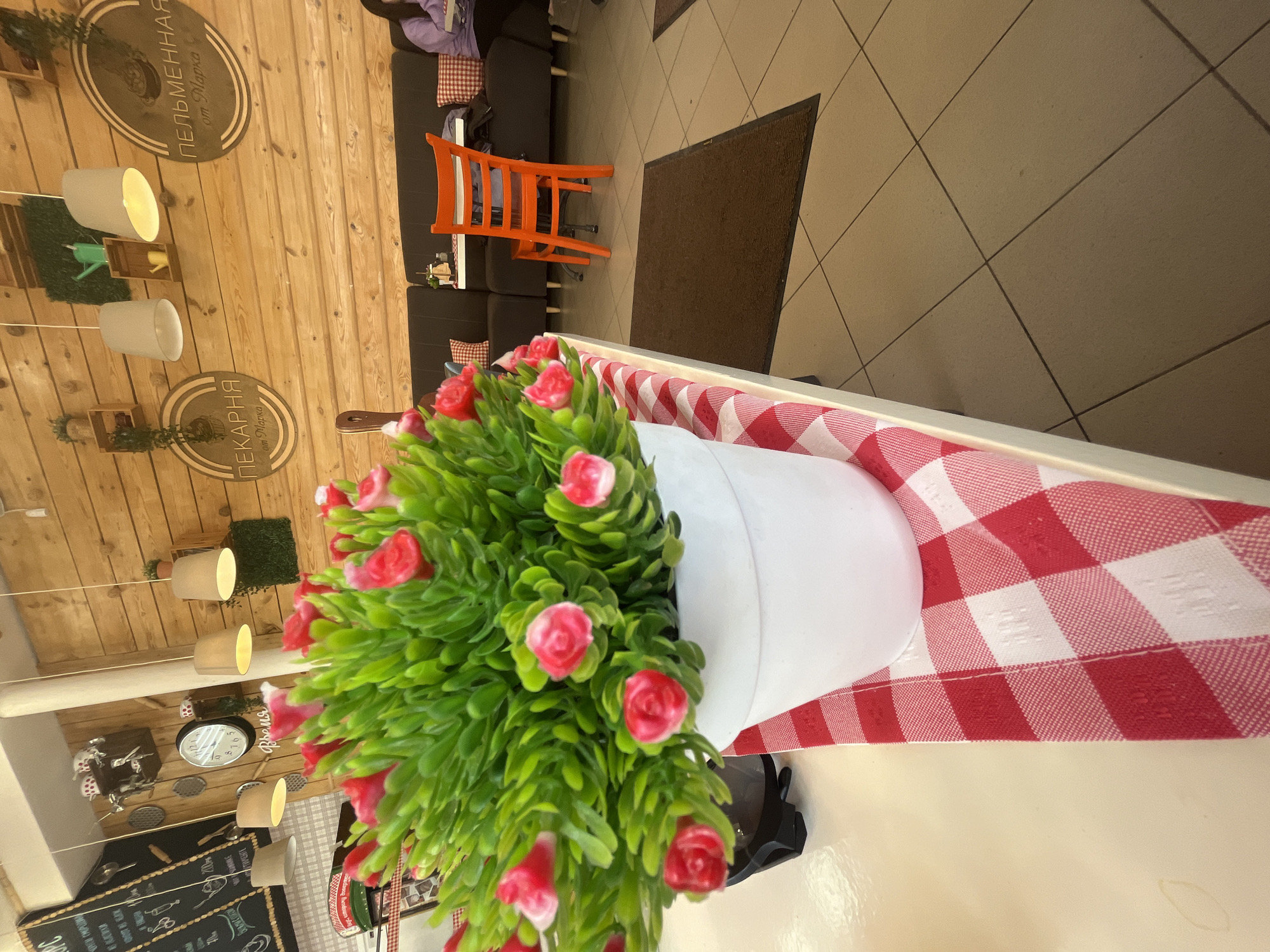 The image, rotated 90 degrees clockwise, presents the interior of a casual cafe or restaurant, likely located in Ulyanovsk, Russia. In the foreground, a large white pot contains an artificial plant with vibrant green foliage and numerous small pink or red flower buds, resembling miniature roses. This pot is positioned on a table covered with a festive red and white checkered tablecloth.
The background reveals the seating and decorative elements of the establishment. The walls are paneled with light-colored wood, creating a warm ambiance. Several hanging light fixtures with off-white shades illuminate the space. Prominently displayed on the wooden wall are two oval signs; one reads "ПЕЛЬМЕННАЯ от Марка" (Dumpling House from Mark), and the other "ПЕКАРНЯ от Марка" (Bakery from Mark), indicating the type of cuisine served. A clock on the wall has the word "Время" (Time) written on it. Further along the wall, a chalkboard menu, though not entirely legible, lists various dishes in Russian.
Seating areas consist of black cushioned sofas and small tables, one of which features a striking orange wooden chair. A person is partially visible, seated on one of the sofas in the background. The floor features light-colored tiles, with a brown doormat marking a transition area. The scene depicts a relaxed and inviting dining environment.