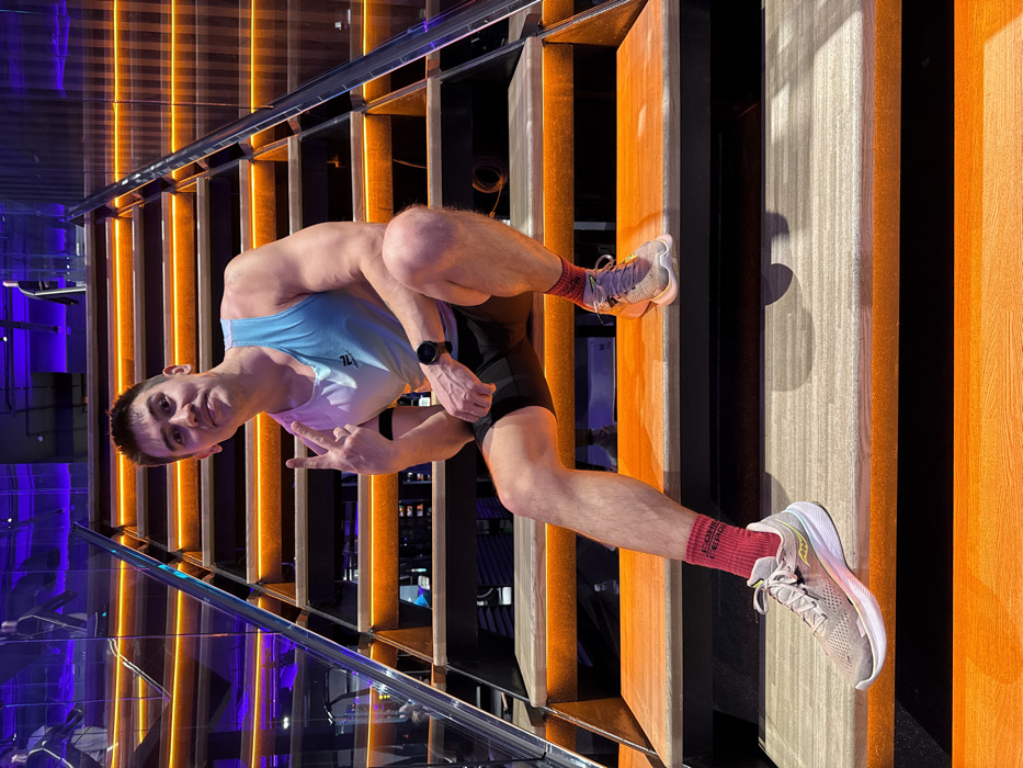 An adult male with short, dark hair and a muscular build is seated on the third step from the bottom of a modern, internally illuminated wooden staircase. He wears a white and light blue athletic tank top, black shorts, red socks with "СОЮЗ СПОРТ" (SOYUZ SPORT) text, and light grey athletic shoes. A black smartwatch is visible on his left wrist, and a black armband is on his left bicep. He is facing forward, looking at the camera with a faint smile, and his left hand is raised in a peace sign. His left leg is extended down to the second step, while his right knee is bent. The staircase features glass balustrades and metal railings, with each step illuminated by an orange LED strip. The background reveals a dark interior space with purple ambient lighting, consistent with a fitness club environment. The presence of "СОЮЗ СПОРТ" on the socks suggests the location is a fitness club within Kazan, Russia, specifically likely the Soyuz Sport facility.