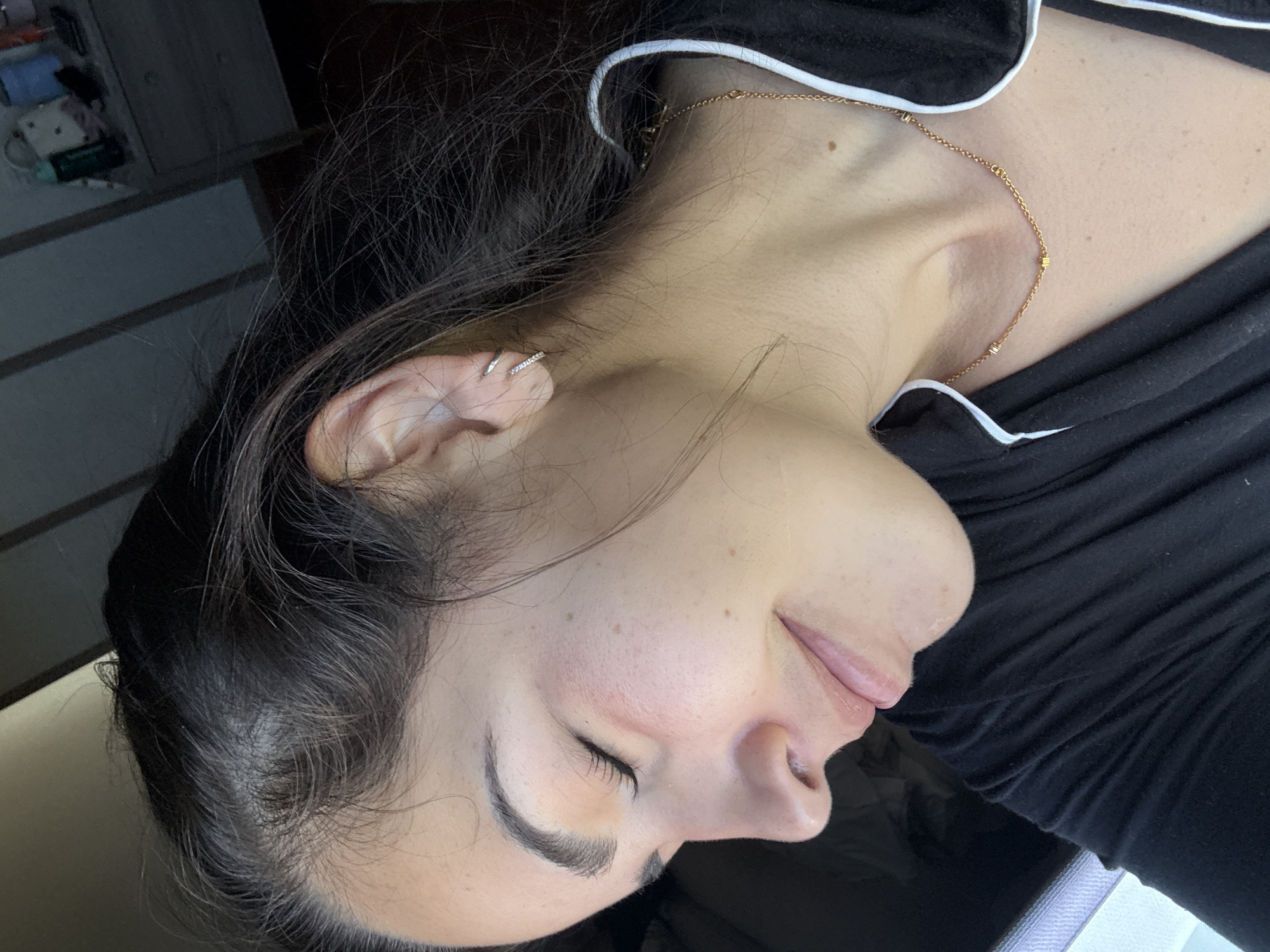 A woman is lying down, facing to the side. She has dark hair, fair skin, and a gold necklace. Her eyes are closed. She is wearing a black top with white trim. There are items on a shelf in the blurred background. The location and city of San Bernardino Tlaxcalancingo, Mexico, are not discernible from the image.
