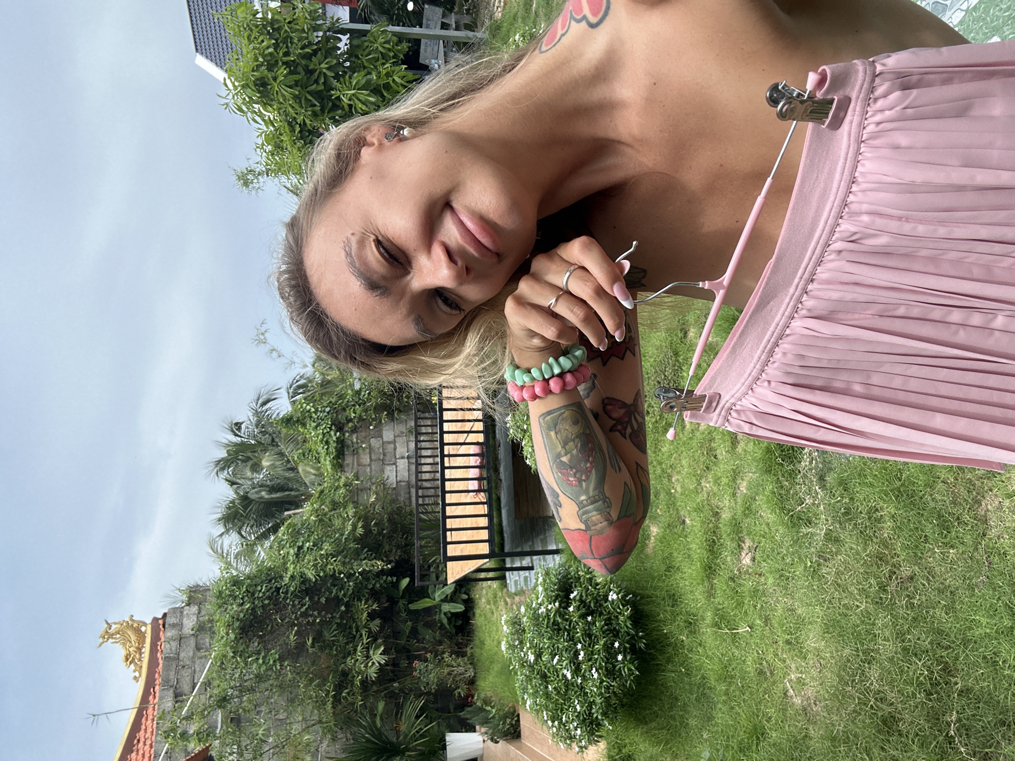 A bright, vertical outdoor portrait features a smiling woman, likely in Mũi Né, Vietnam. She has blonde hair and a tanned complexion, with one eye playfully squinted as she looks towards the camera. She is wearing earrings and has several rings on her fingers, along with two beaded bracelets in shades of turquoise/green and pink on her left wrist. Her left arm and upper shoulder are visibly adorned with colorful tattoos, including a prominent design of a ship in a bottle and a red rose on her forearm, and a floral pattern on her shoulder.
The woman is holding a pink pleated skirt on a matching pink clothes hanger, extending it towards the lower right of the frame. The background reveals a lush, tropical environment typical of Mũi Né, with abundant green grass and dense foliage, including palm trees and bushes with small white flowers. Architectural elements are visible, including a dark-tiled roof of a building with an intricate golden ornament, possibly a decorative feature of a local temple or traditional structure. A set of wooden stairs with metal railings can be seen amidst the greenery. The scene is brightly lit by natural daylight, indicating clear and sunny weather. No visible text is present.