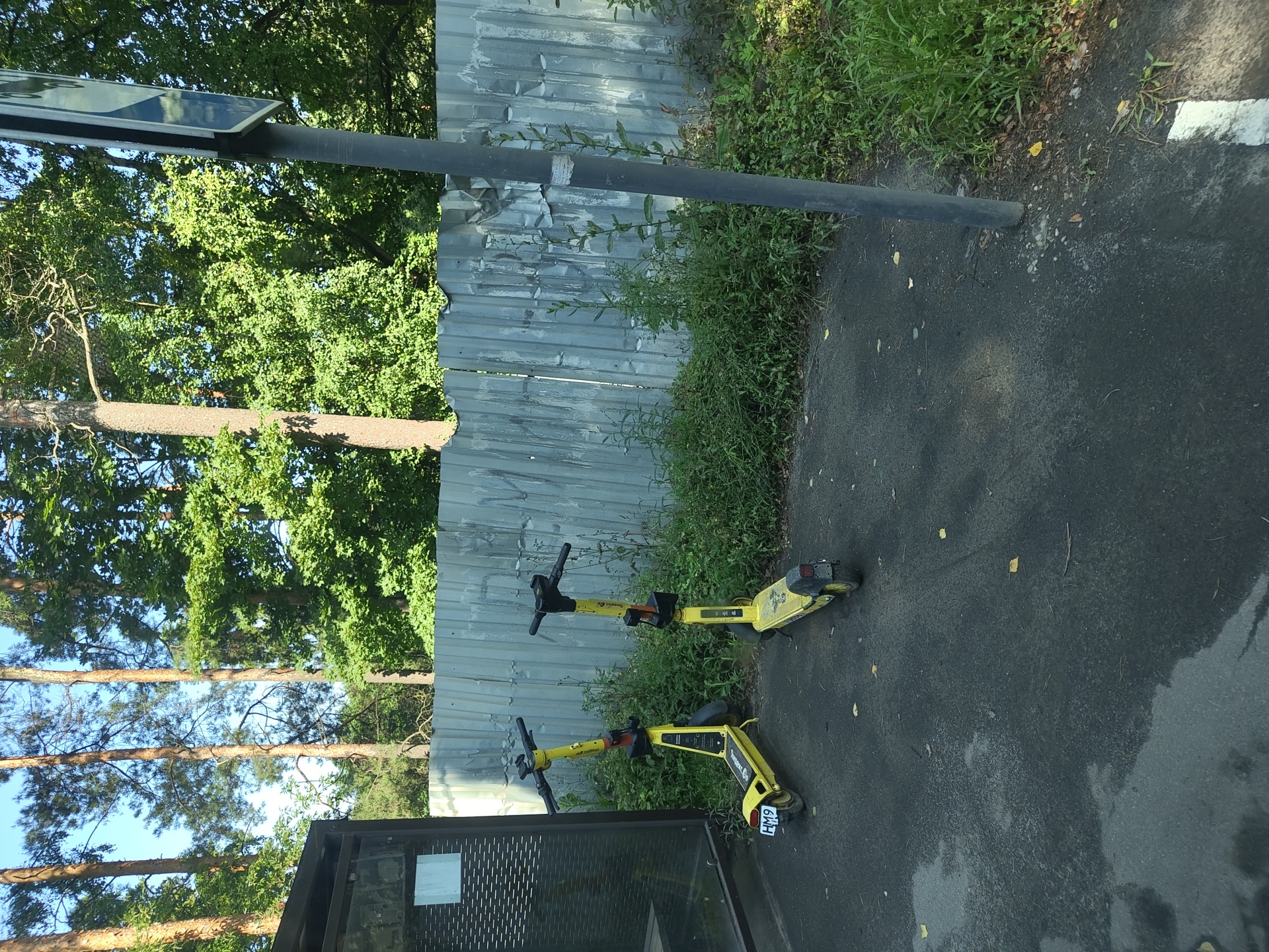 Two yellow electric scooters are parked on a paved surface next to a corrugated metal fence and a bus stop shelter. The scooter on the left has a license plate that reads "HM9 714". Green foliage and trees are visible behind the fence, and a pole with a sign is on the right side of the image. The ground in front of the scooters is dark asphalt with some patches of lighter color and a few scattered leaves. This scene appears to be an outdoor public space in Ramenskoye, Russia.