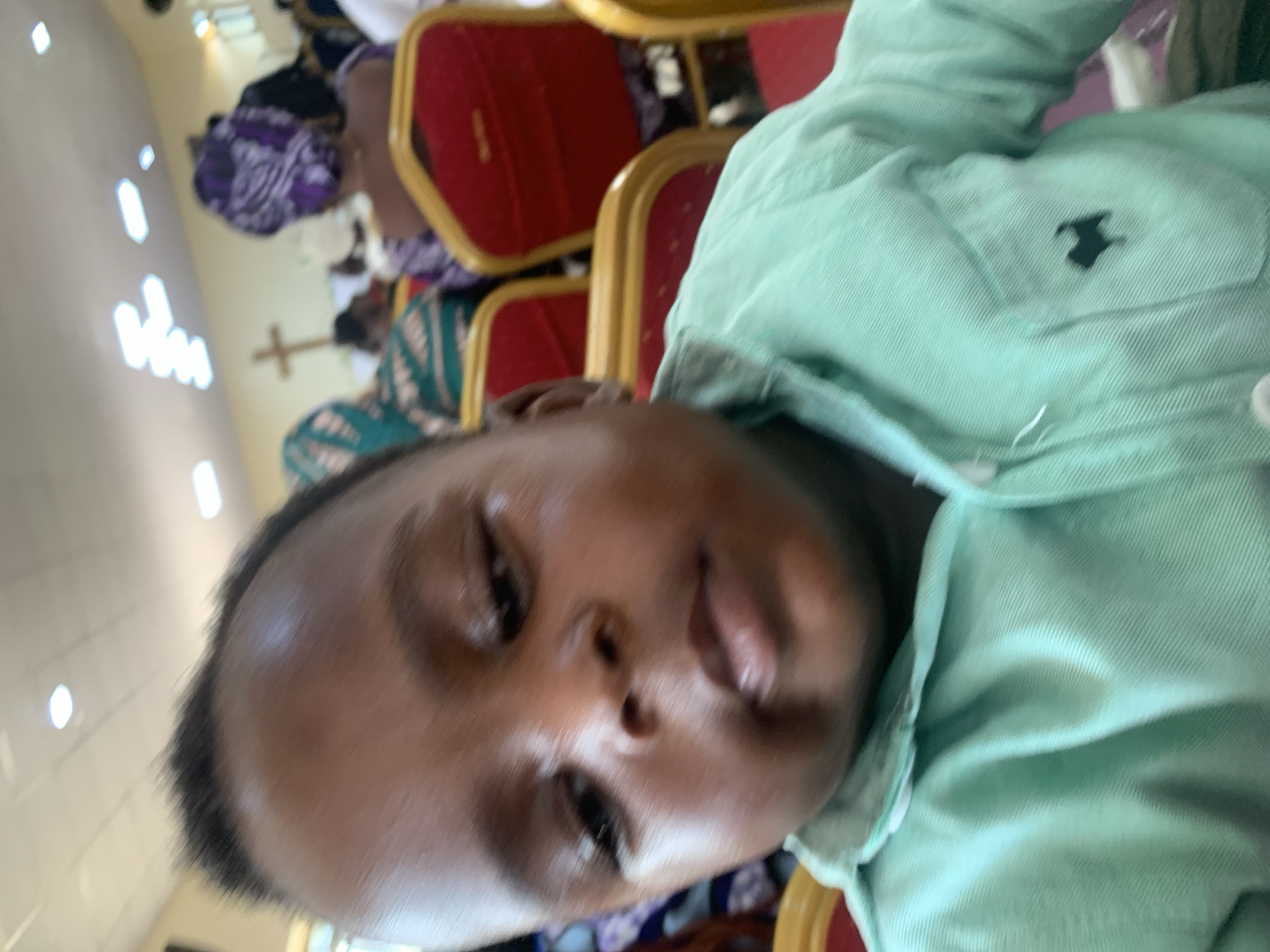 An extreme close-up shot shows the face of a young Black child from the chest up, looking slightly downward and to the left. The child is wearing a light mint green collared shirt with a small black silhouette of a dog embroidered on the left breast pocket. The background is blurred but appears to be a church setting, with rows of red and gold chairs and several adults wearing headscarves. A wooden cross is visible on the wall in the background. The lighting is bright, possibly from overhead fluorescent lights. The overall impression is of a candid snapshot taken during a service or gathering.