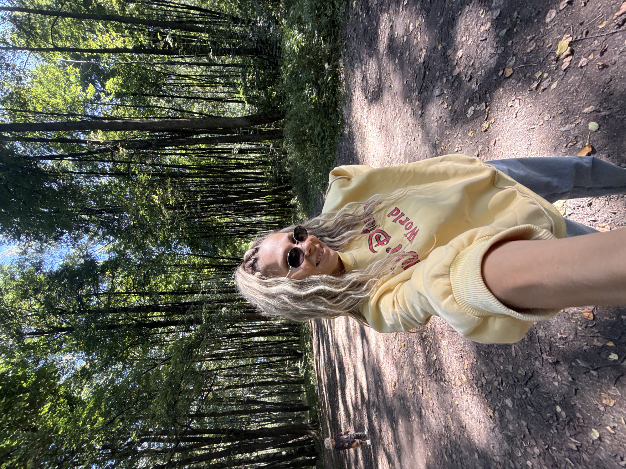 In Rusanovo, Russia, a woman with blonde braided hair and sunglasses is taking a selfie in a sunlit forest. She is wearing a yellow sweatshirt with red lettering that says "Disney World." Her right arm is extended towards the camera. In the background, a young child wearing a patterned outfit and a hat is walking away from the camera on a dirt path. The path is lined with tall trees with green leaves, and sunlight filters through the canopy, casting shadows on the ground. The ground is covered with dirt and fallen leaves.
