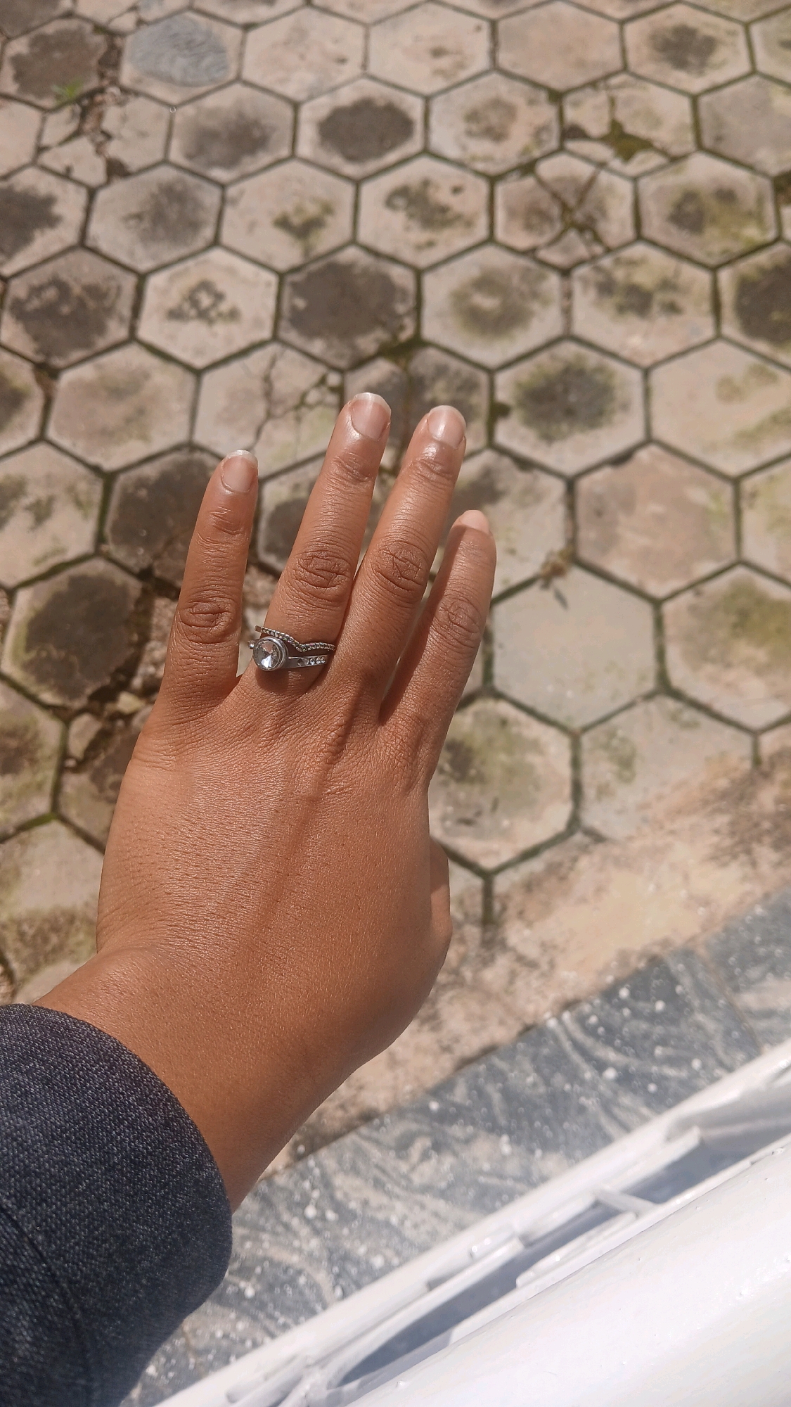 A person's hand is held up, displaying a silver ring with a clear stone and a band of smaller stones. The hand is positioned over a textured, hexagonal-tiled surface. The tiles are a light beige color with darker, irregular patches of discoloration and some moss. The background shows more of the same tiling pattern. A dark gray sleeve of clothing is visible on the lower left of the frame. The scene appears to be outdoors, possibly in a courtyard or paved area.