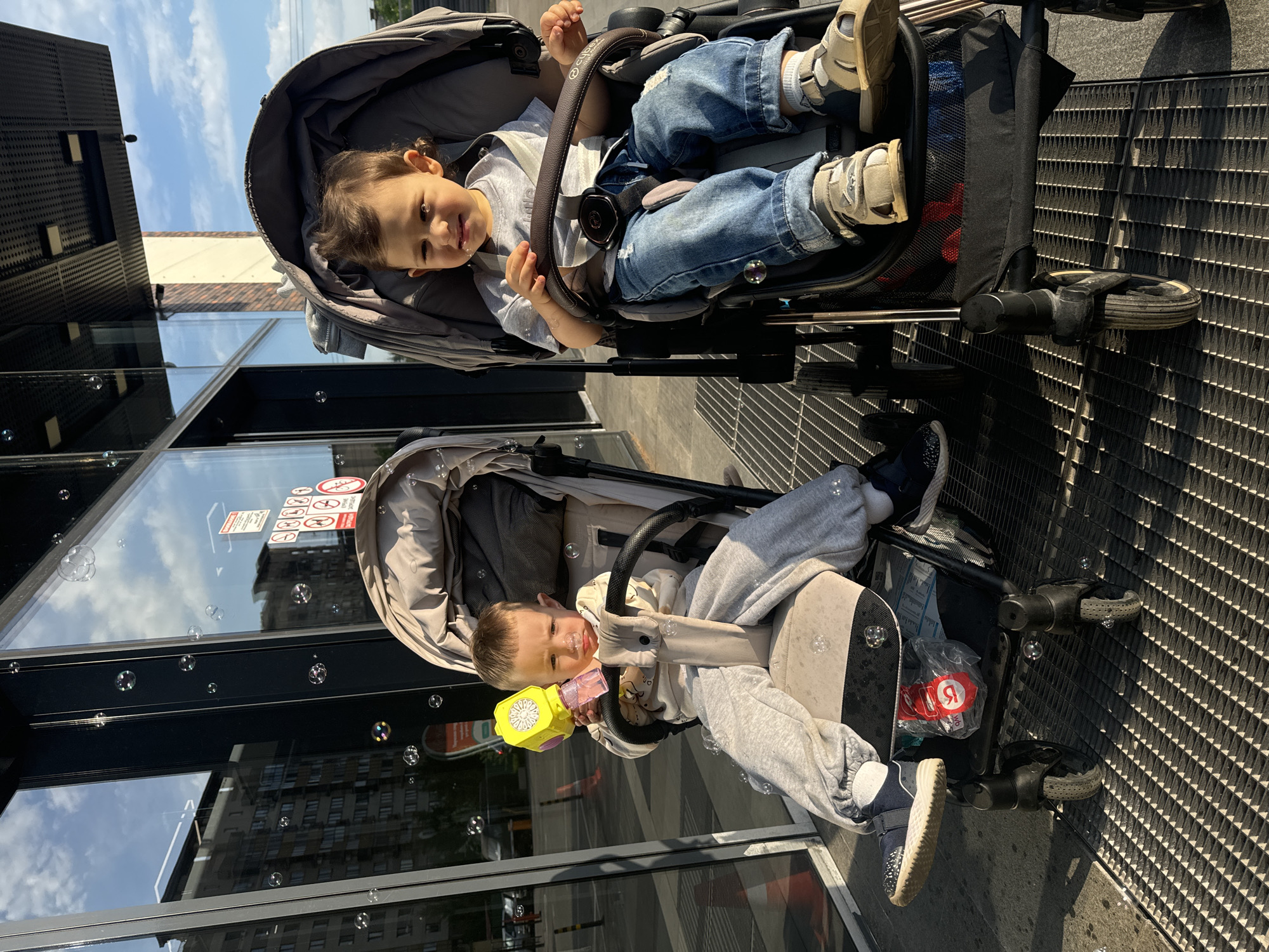 Two young children are seated in strollers on a metal grated surface, likely in Unknown, Russia, based on the Cyrillic script visible. The child in the top stroller is wearing jeans, a light-colored shirt, and sandals. The child in the bottom stroller is wearing a grey tracksuit and sneakers, and is holding a yellow and pink toy bubble gun. Numerous bubbles are visible around the strollers. The background shows a modern building with glass windows reflecting the sky and clouds. Stickers and signs are visible on the glass.