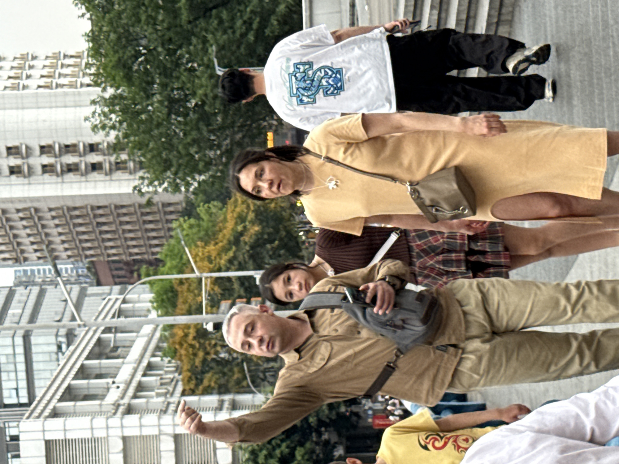 The image, rotated for standard viewing, depicts a group of people on an urban pedestrian walkway in Yuzhong District, China. In the foreground, a man with short, gray hair, wearing a tan shirt and pants with a gray sling bag, gestures with his right hand. Behind him, a young girl with dark hair and a striped top looks towards the camera. Further back, a woman in a yellow dress has a small gray purse hanging from her shoulder. Behind her and slightly to the left is a man in a white t-shirt with a graphic design, black pants, and sneakers. Buildings are visible in the background.