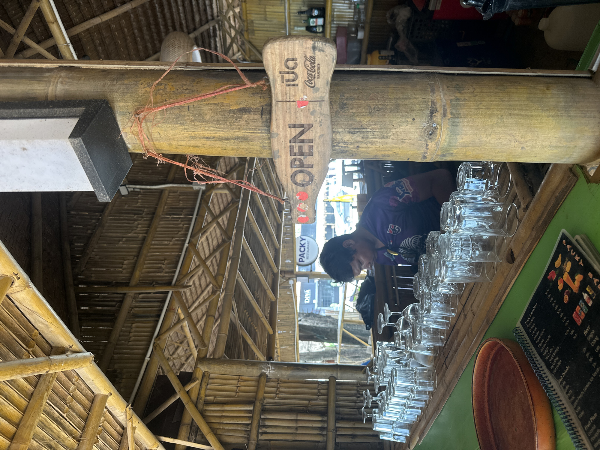 The image shows an indoor bar area with a rustic, bamboo-themed decor. A man wearing a purple shirt is standing behind a bar counter covered with numerous clean drinking glasses. Above him, a wooden sign shaped like a Coca-Cola bottle reads "OPEN" in English and Thai script. The bar appears to be located in a tropical or beachside setting, as suggested by the open-air structure and the presence of palm trees visible through the windows in the background. A menu is also visible on the green counter in the foreground, listing various drinks and food items. The overall ambiance is casual and inviting, suggesting a bar or restaurant establishment.