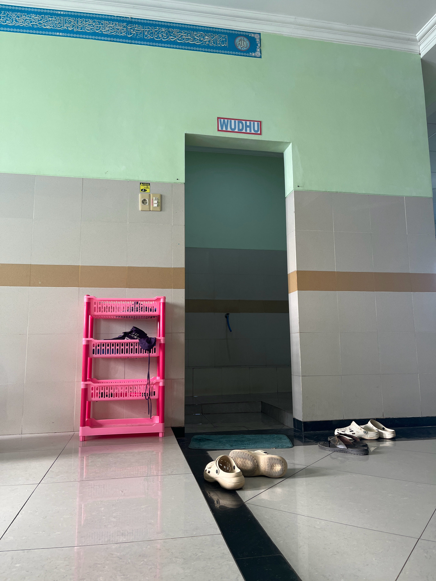 The image shows an interior scene, likely a mosque or public facility, with a light green painted wall at the top and tiled walls below. A blue Arabic inscription is visible on the top portion of the wall. A red sign with white lettering reading "WUDHU" is affixed to the wall above an open doorway. To the left of the doorway is a beige electrical outlet with a switch. A bright pink three-tiered plastic shelf unit stands against the tiled wall, holding a dark purple garment. The floor is tiled with large light grey tiles with darker grout lines, and a reflective surface indicates a polished finish. Several pairs of sandals are arranged on the floor near the doorway. In the foreground, two beige clogs are visible. The doorway leads into a darker tiled room. The scene is located in Kecamatan Pati, Indonesia.